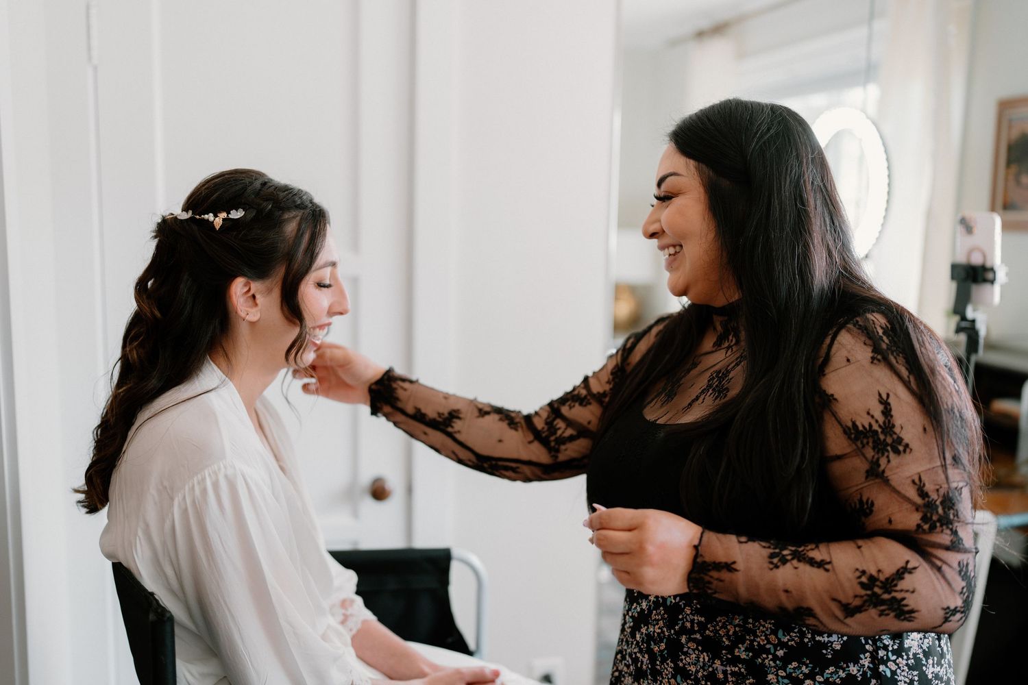 Smiling makeup artist in black lace dress applying earring to bride in white robe during wedding preparations.