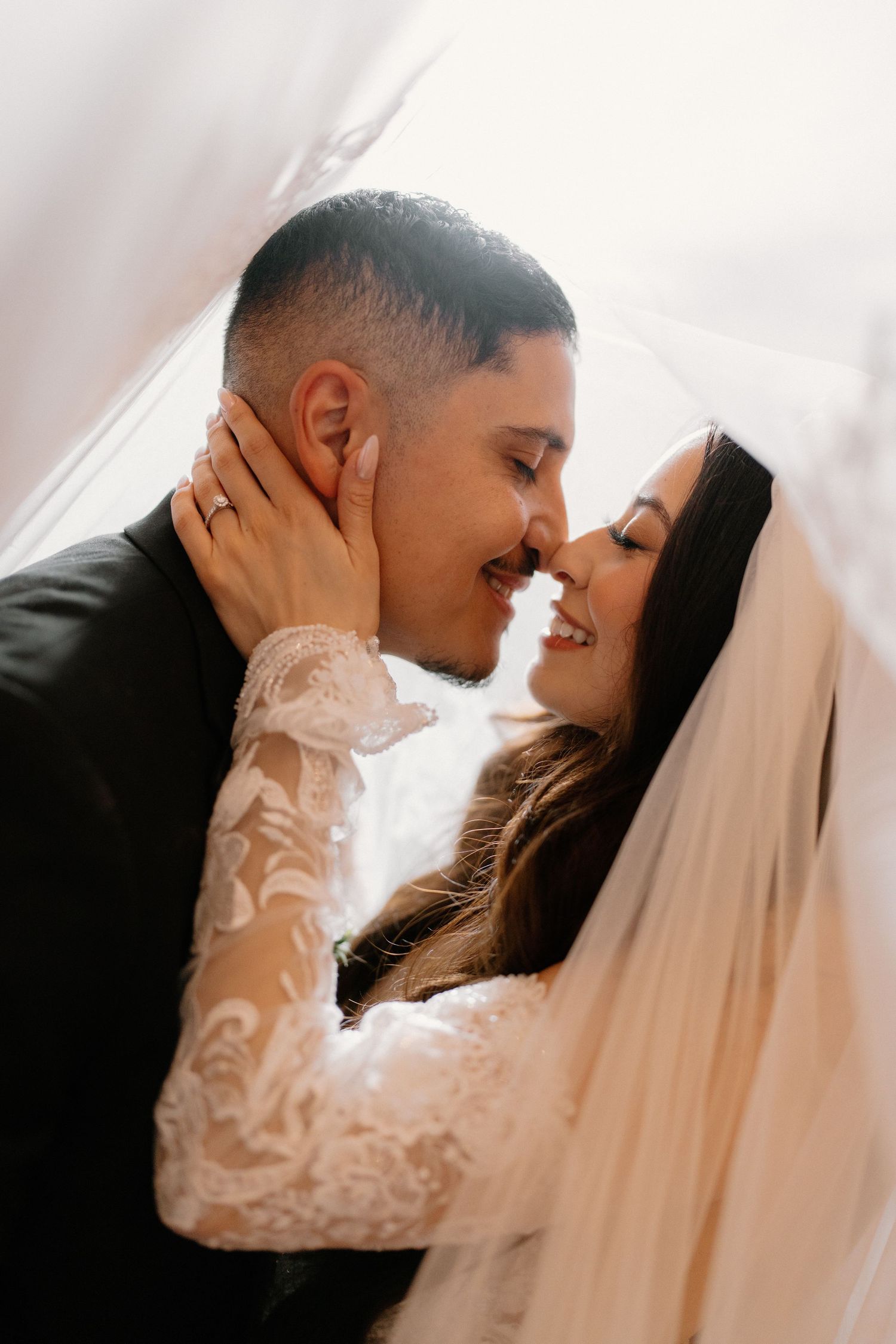 Intimate close-up of smiling bride and groom about to kiss beneath flowing wedding veil.