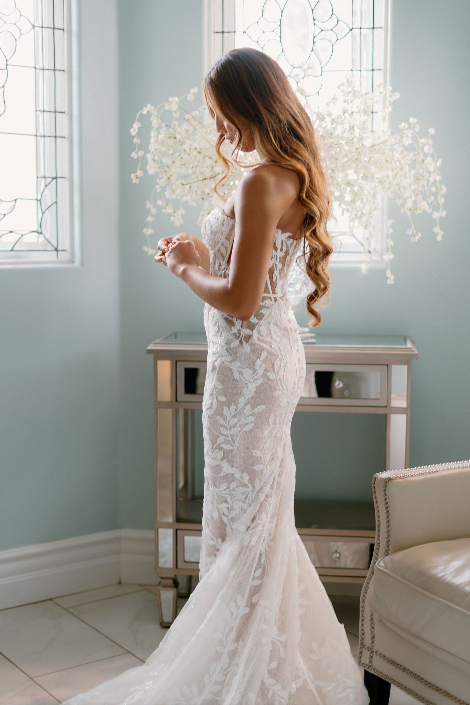 Bride in stunning backless lace mermaid wedding gown stands by mirrored vanity in soft blue bridal suite.