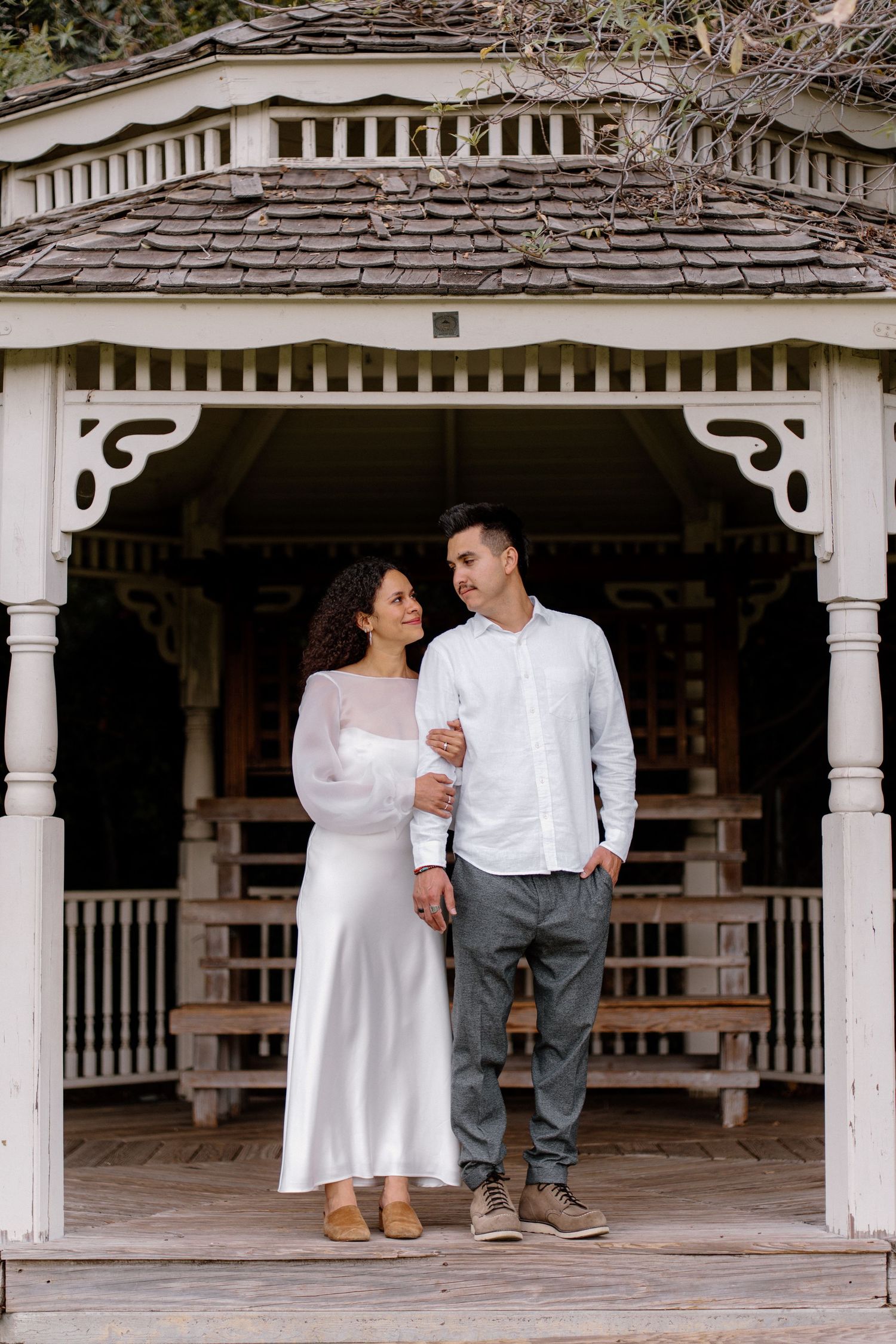 Engaged couple standing together in a white gazebo, woman in white satin dress and man in white shirt.