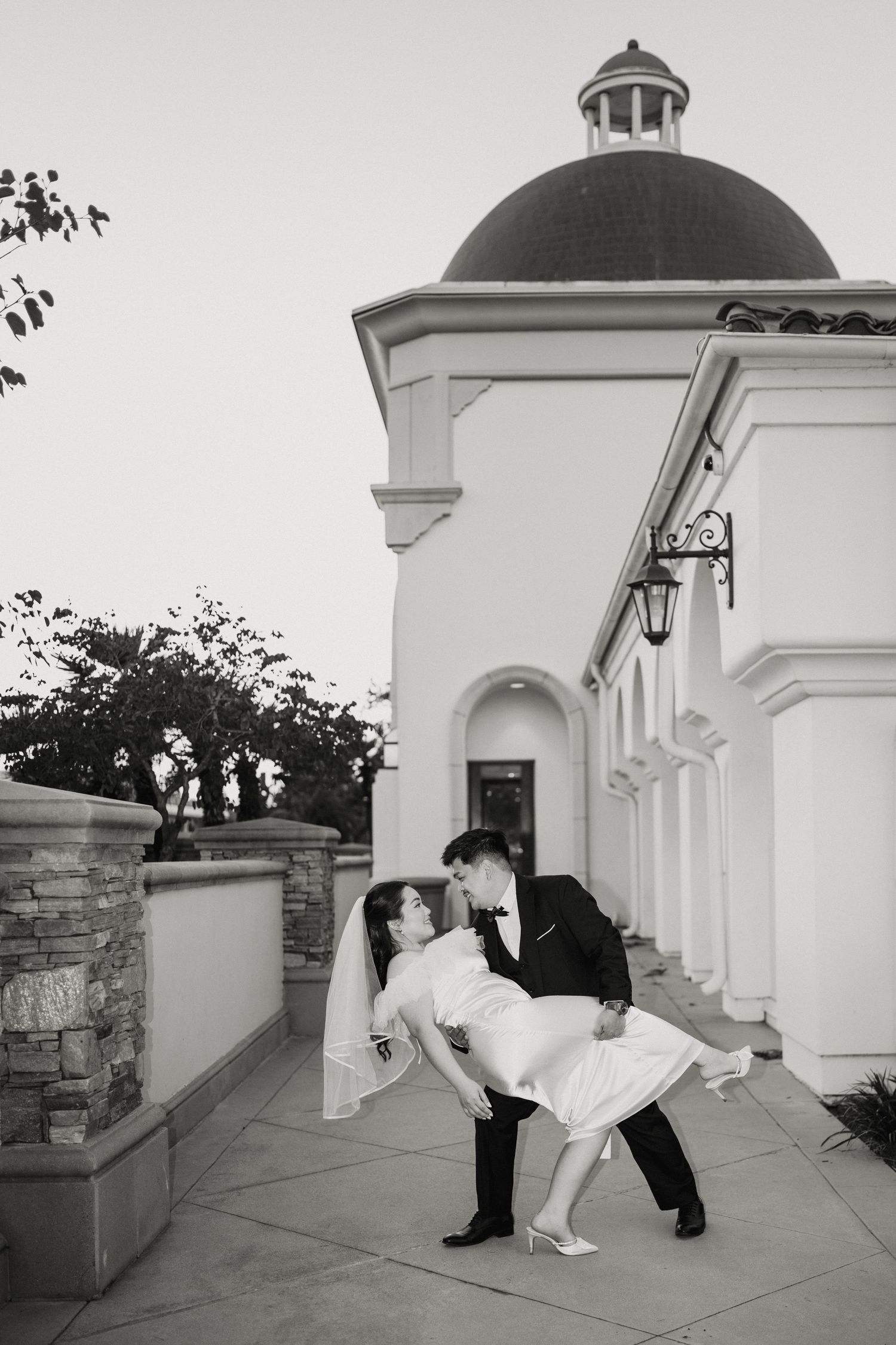 Groom dips bride for a kiss outside a white building with a dome during their wedding day.