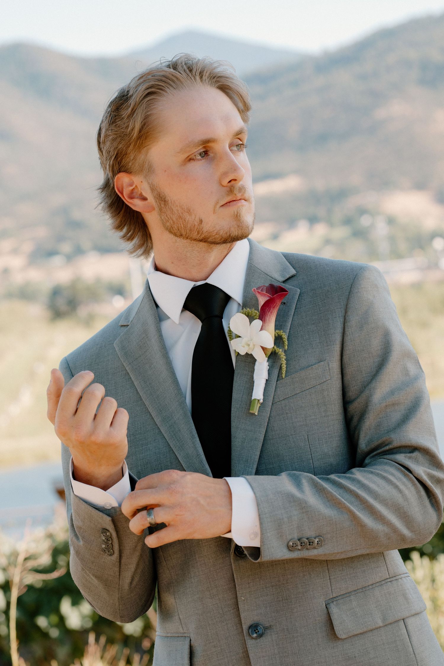Groom in gray suit with black tie and boutonniere adjusting cuffs outdoors with mountain backdrop.