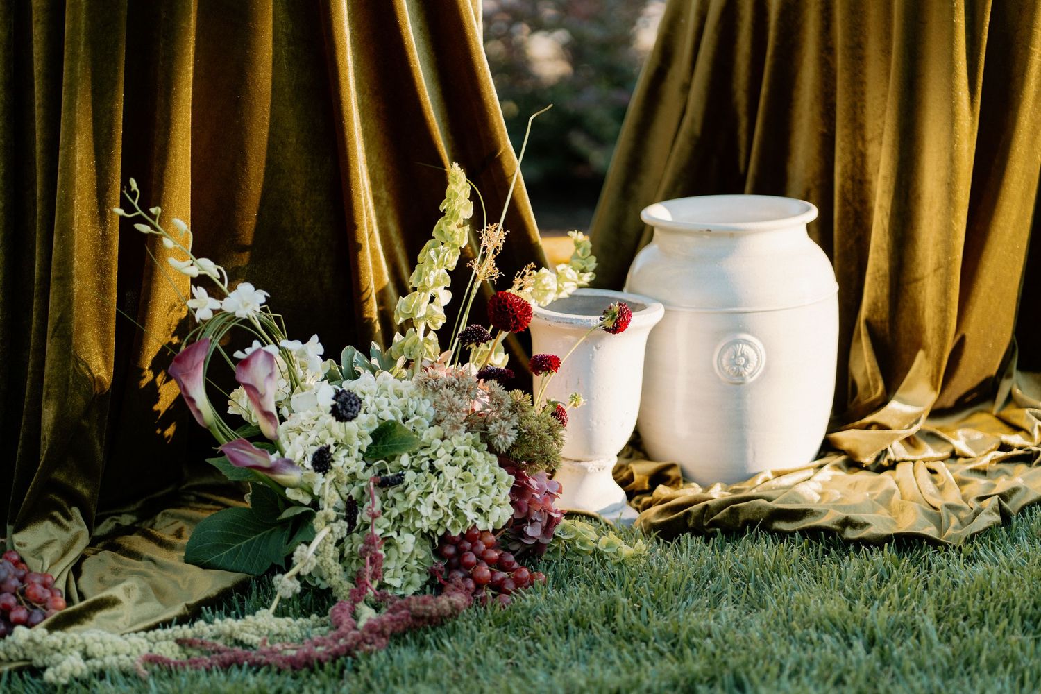 White ceramic urns and wildflower bouquet arranged against golden velvet draping on green grass.