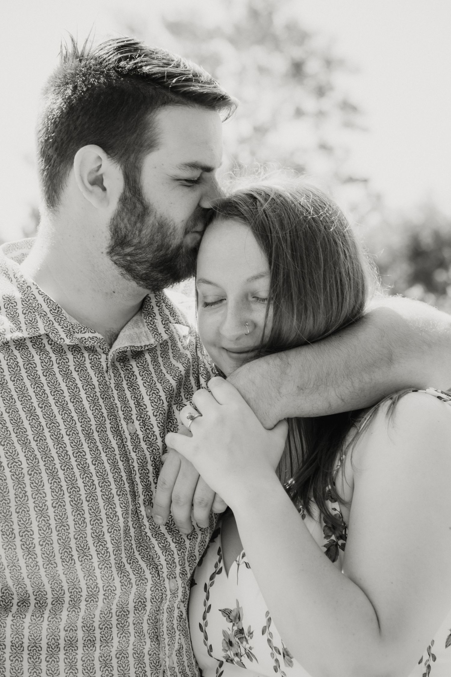 Black and white photo of bearded man kissing woman on head while holding hands tenderly.