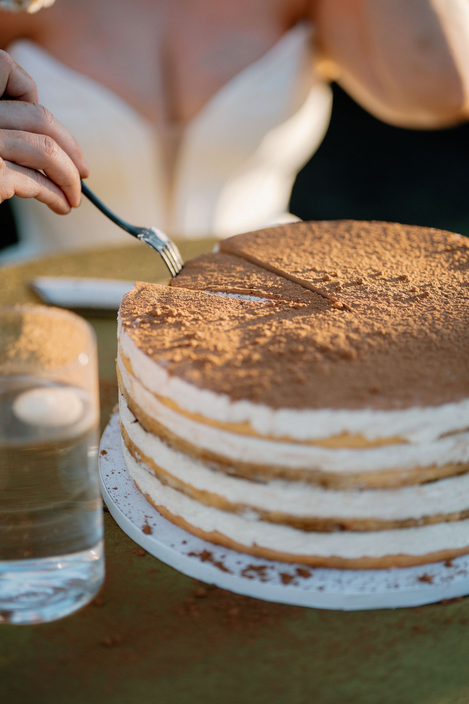 Bride cutting a layered tiramisu wedding cake dusted with cocoa powder at outdoor reception.
