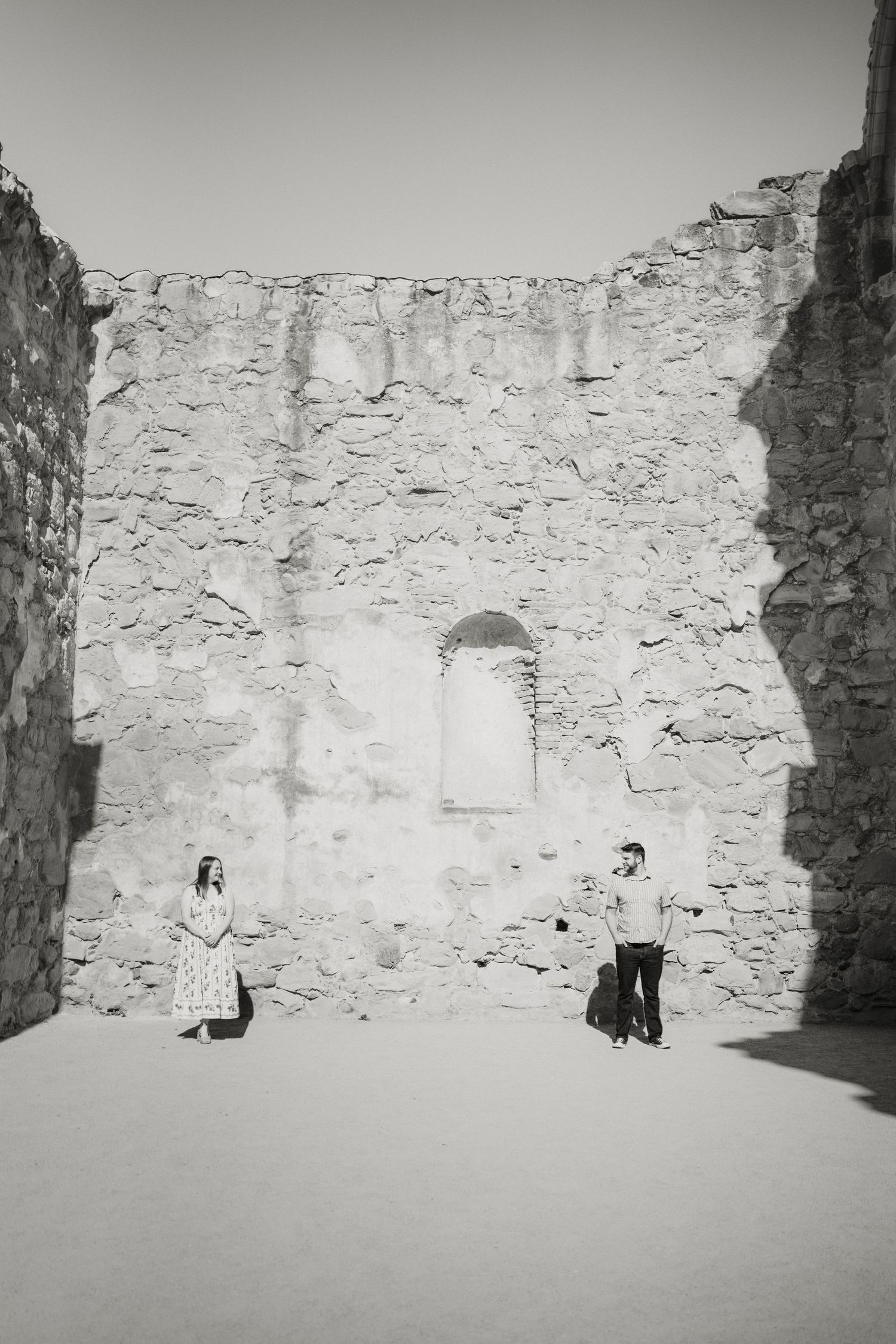 Black and white photo of couple standing apart in ancient stone ruins with dramatic shadows on walls.
