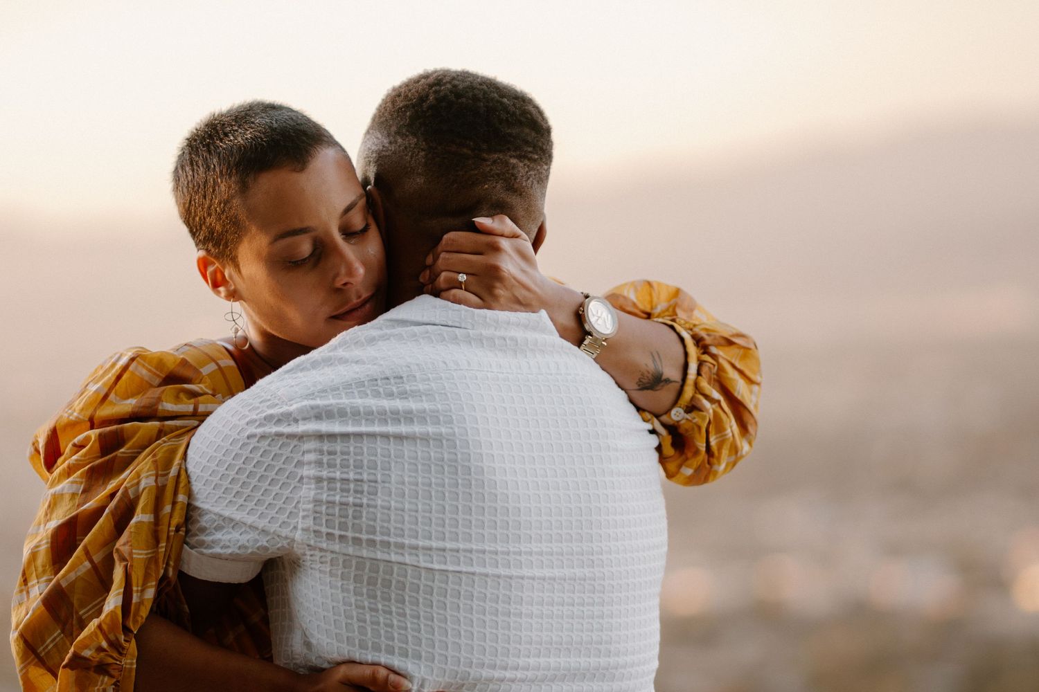 A couple shares a tender embrace at sunset with a scenic cityscape in the background.