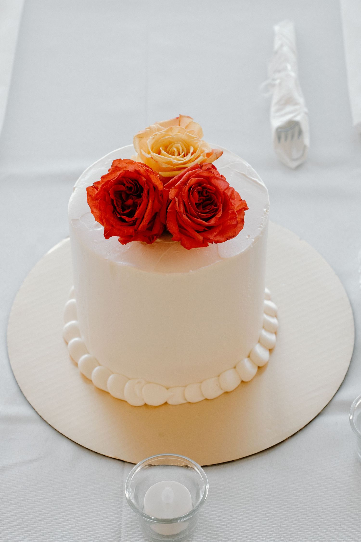 Elegant white wedding cake topped with red and orange roses on white tablecloth.