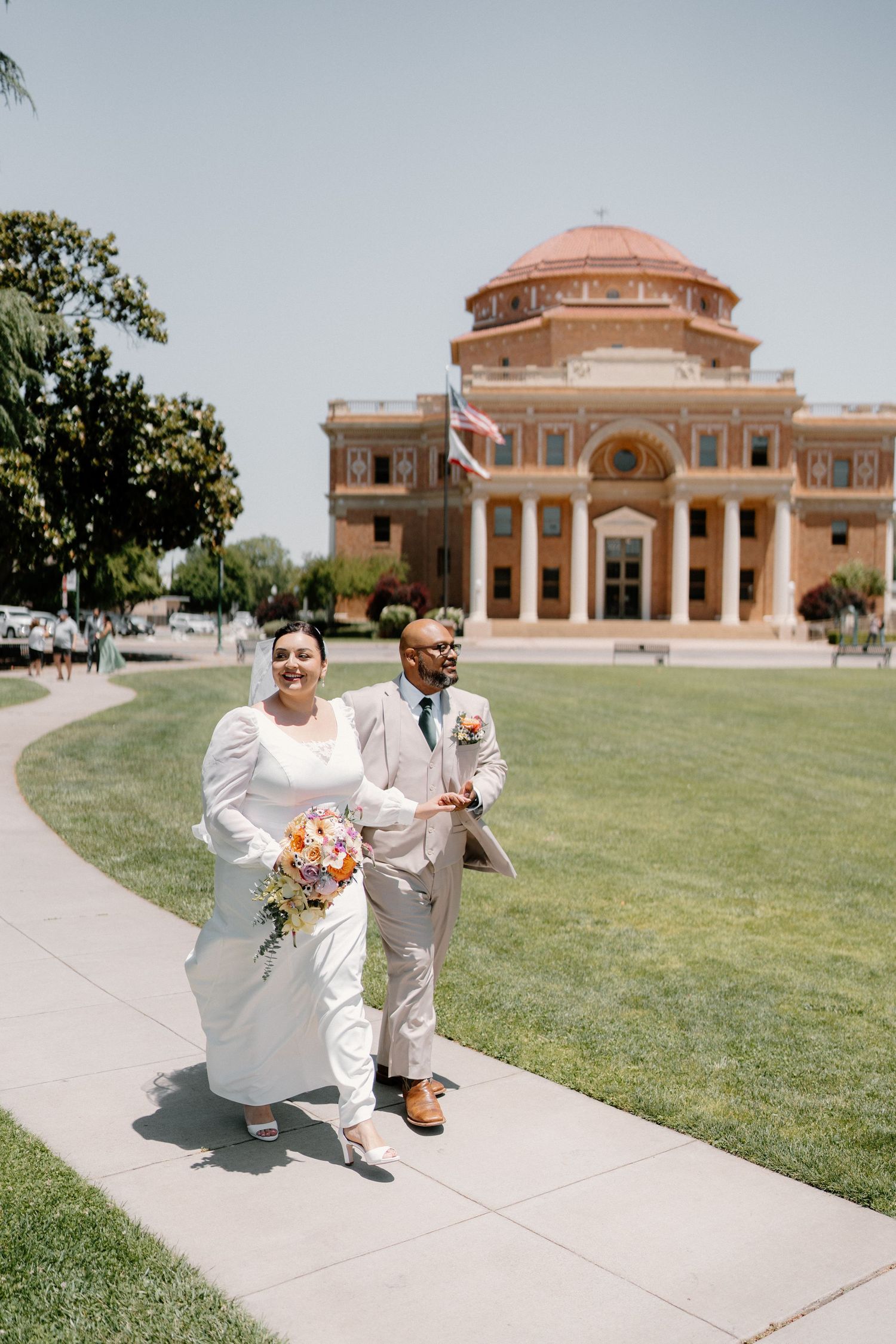 Interracial couple walking from historic domed building after wedding, bride holding colorful bouquet.