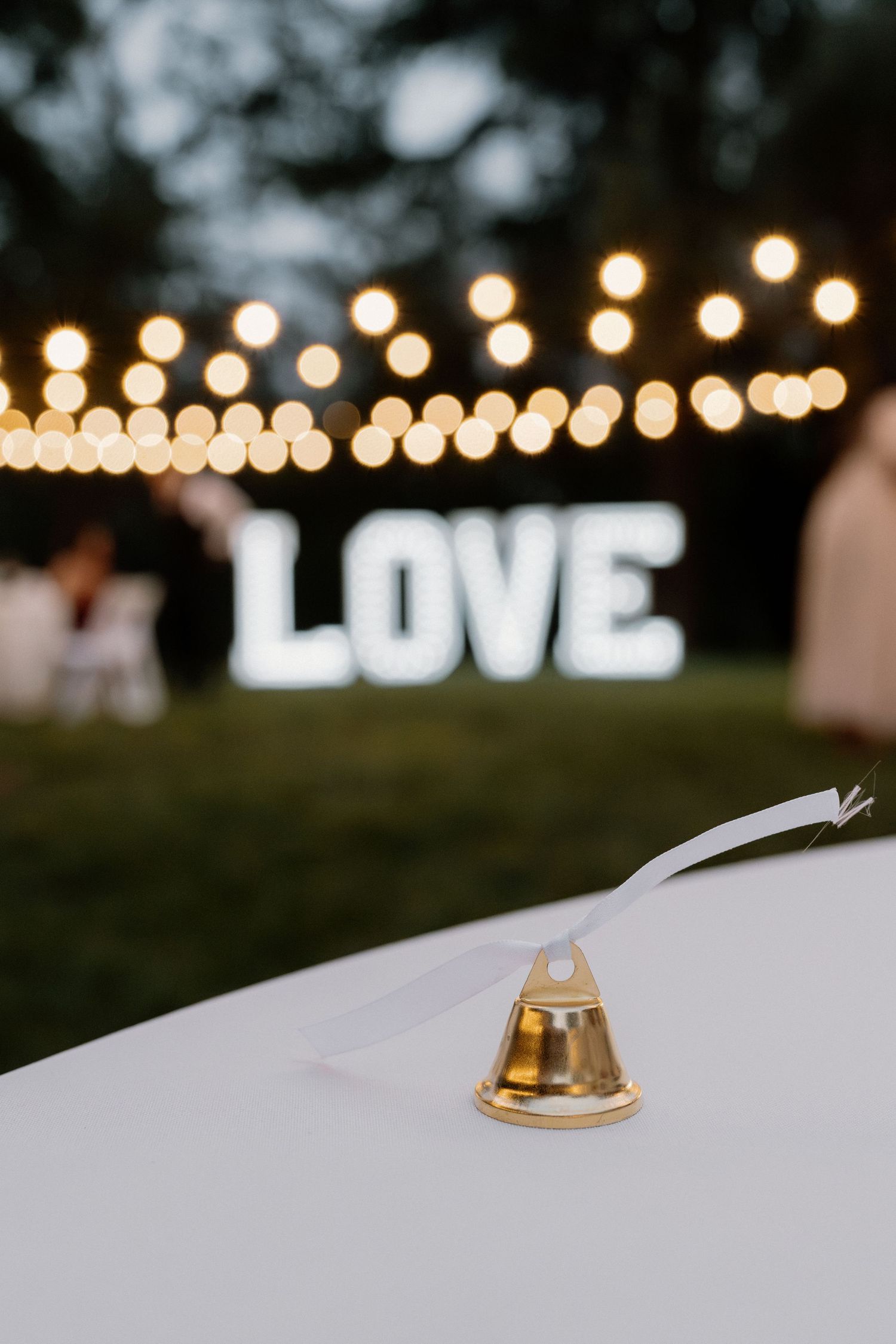 Gold bell favor sits on white tablecloth with illuminated LOVE sign and string lights in background.