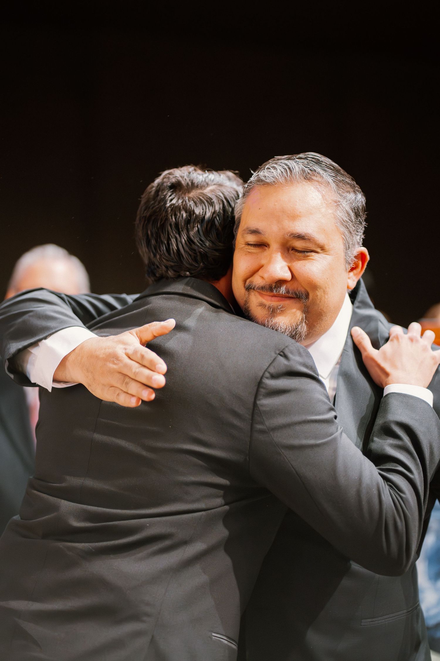 Two men in suits share a warm emotional embrace at a formal wedding ceremony event.