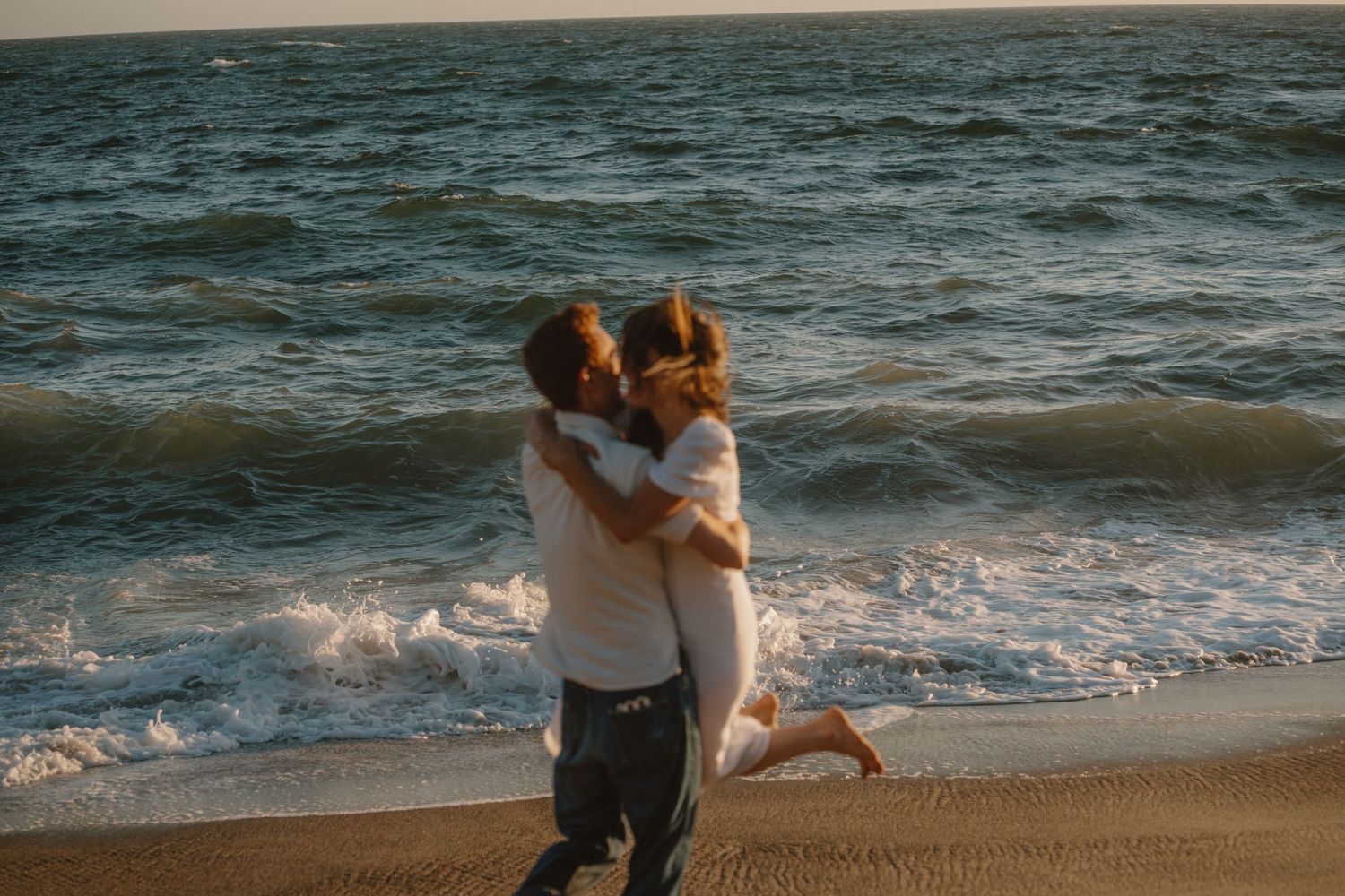 Joyful couple embracing and kissing on sandy beach with ocean waves crashing behind them.