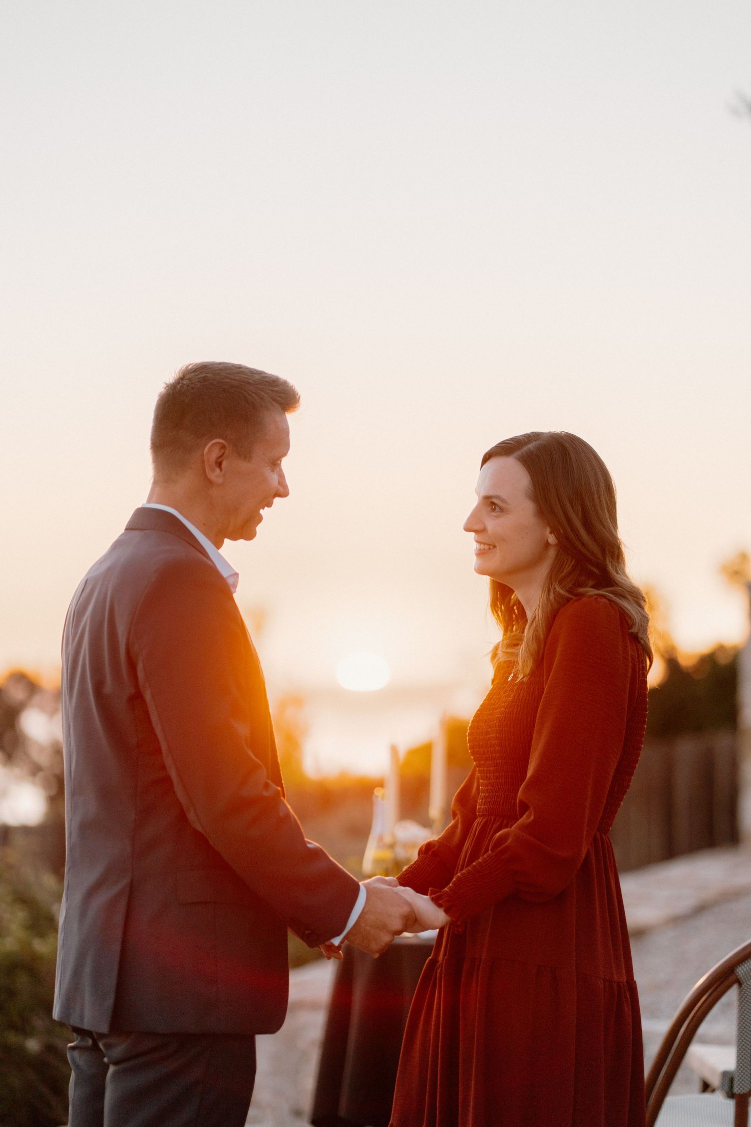 Couple holds hands facing each other bathed in golden sunset light outdoors.