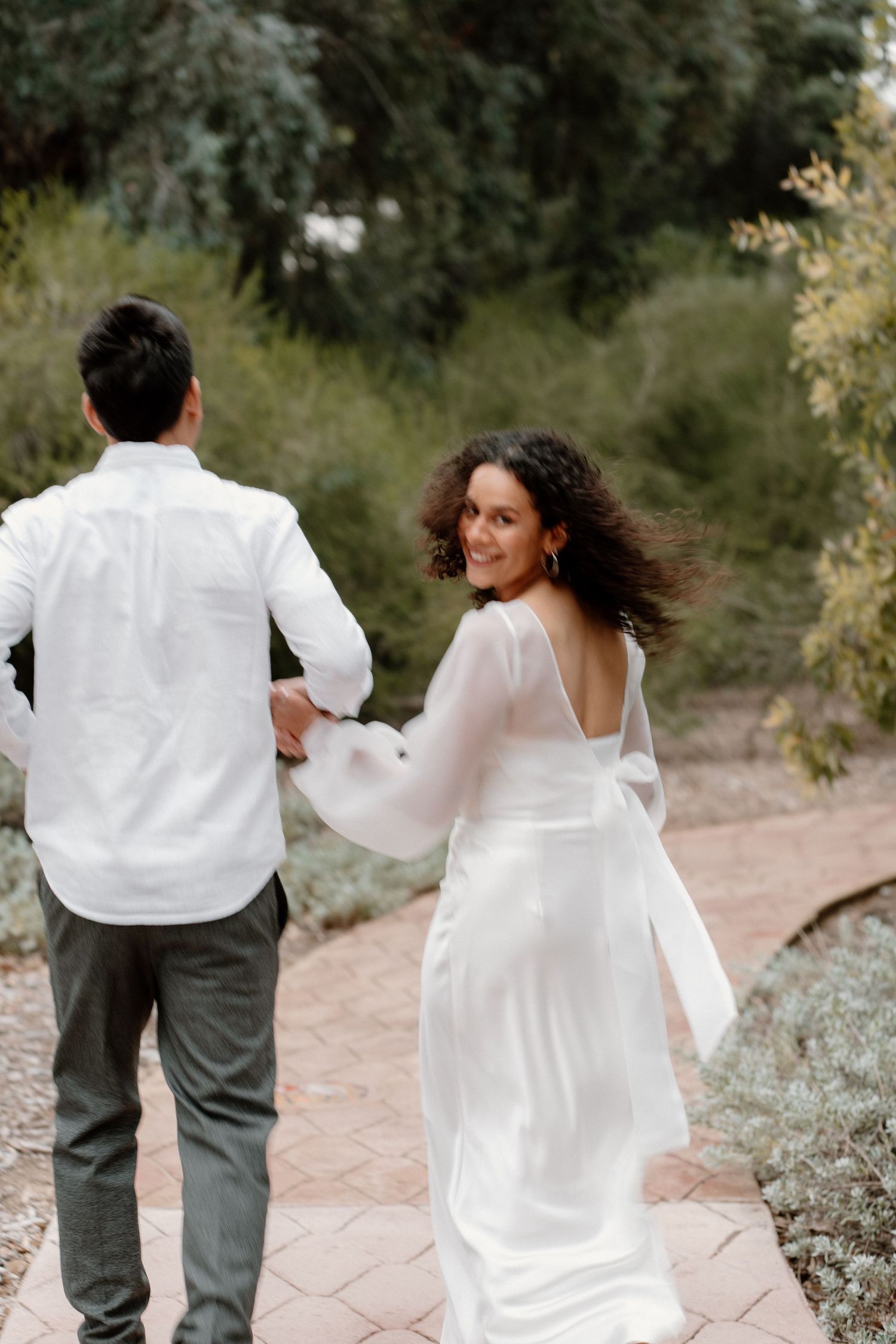 Bride in white backless dress smiling over her shoulder while holding groom's hand on garden path.