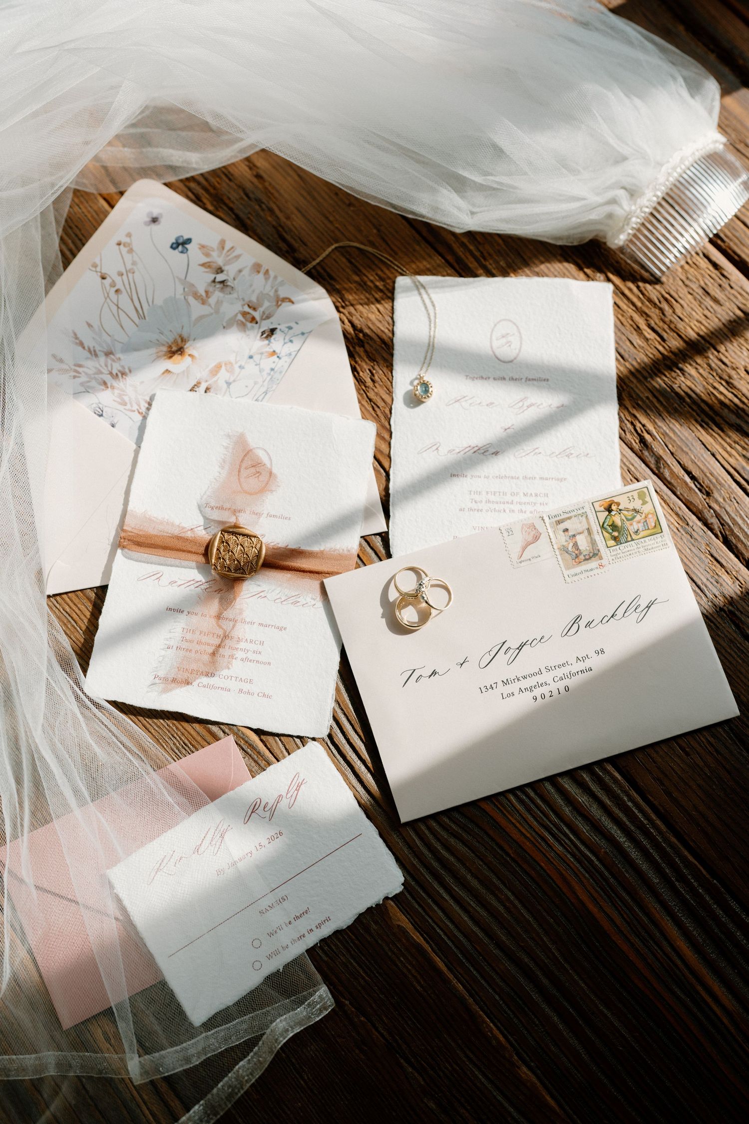 Romantic wedding invitation suite with floral details, wax seal, ribbon, and rings on rustic wood surface.