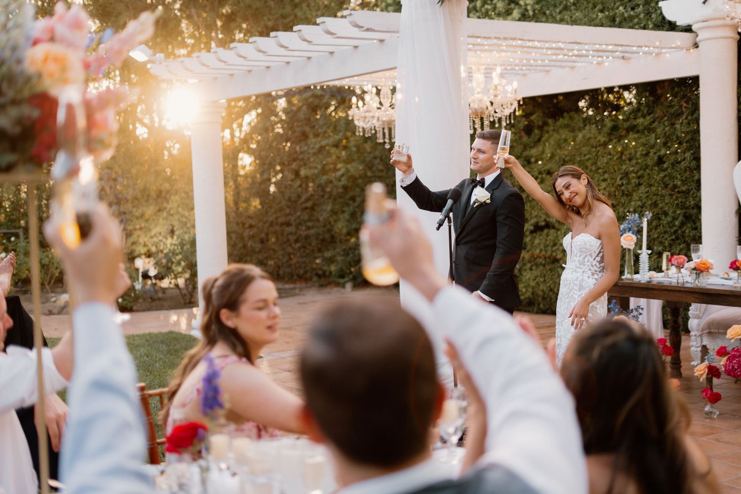 Bride and groom raise glasses during golden hour outdoor wedding reception toast with cheering guests.