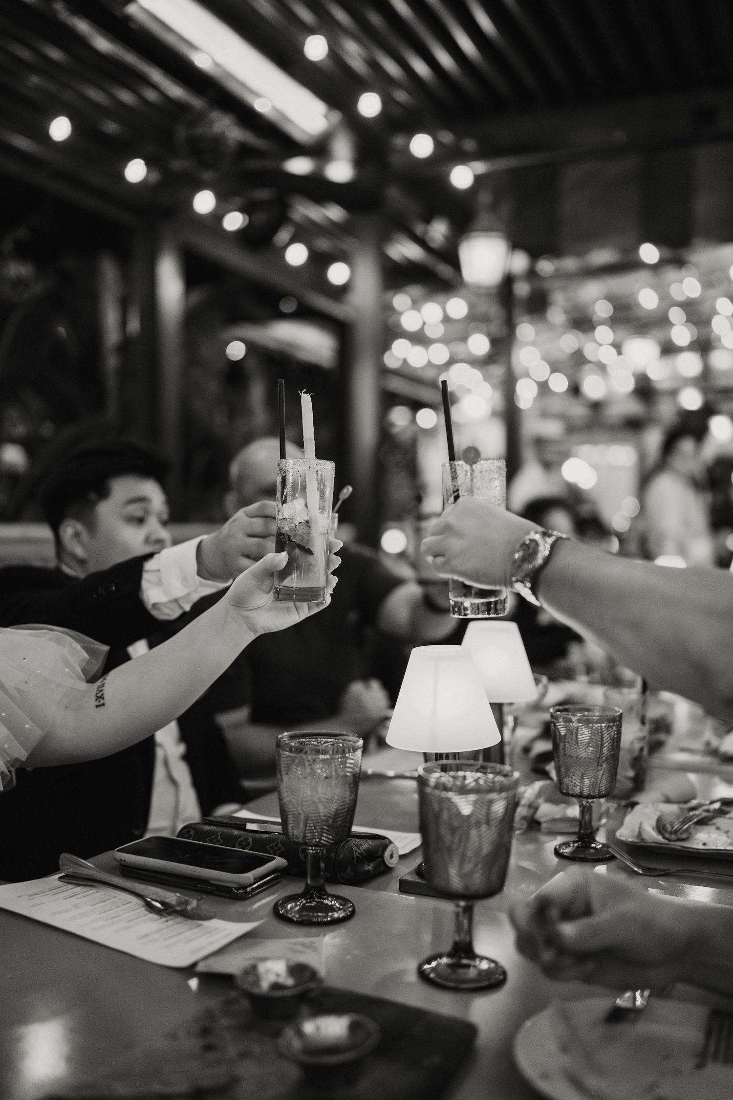Wedding guests clink cocktail glasses in toast at reception with bokeh string lights, black and white.