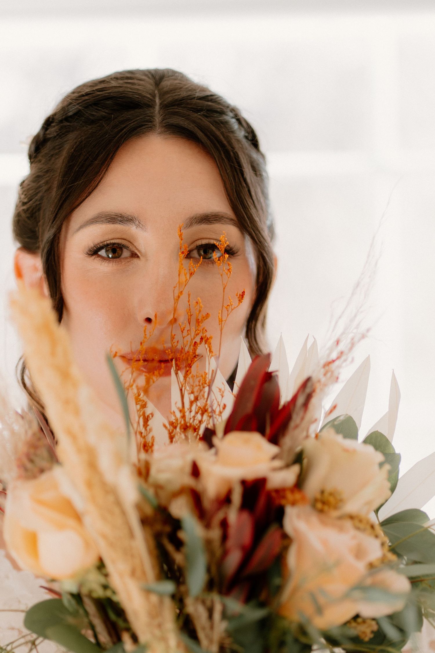 A bride with golden eye makeup peers over a rustic autumn wedding bouquet.