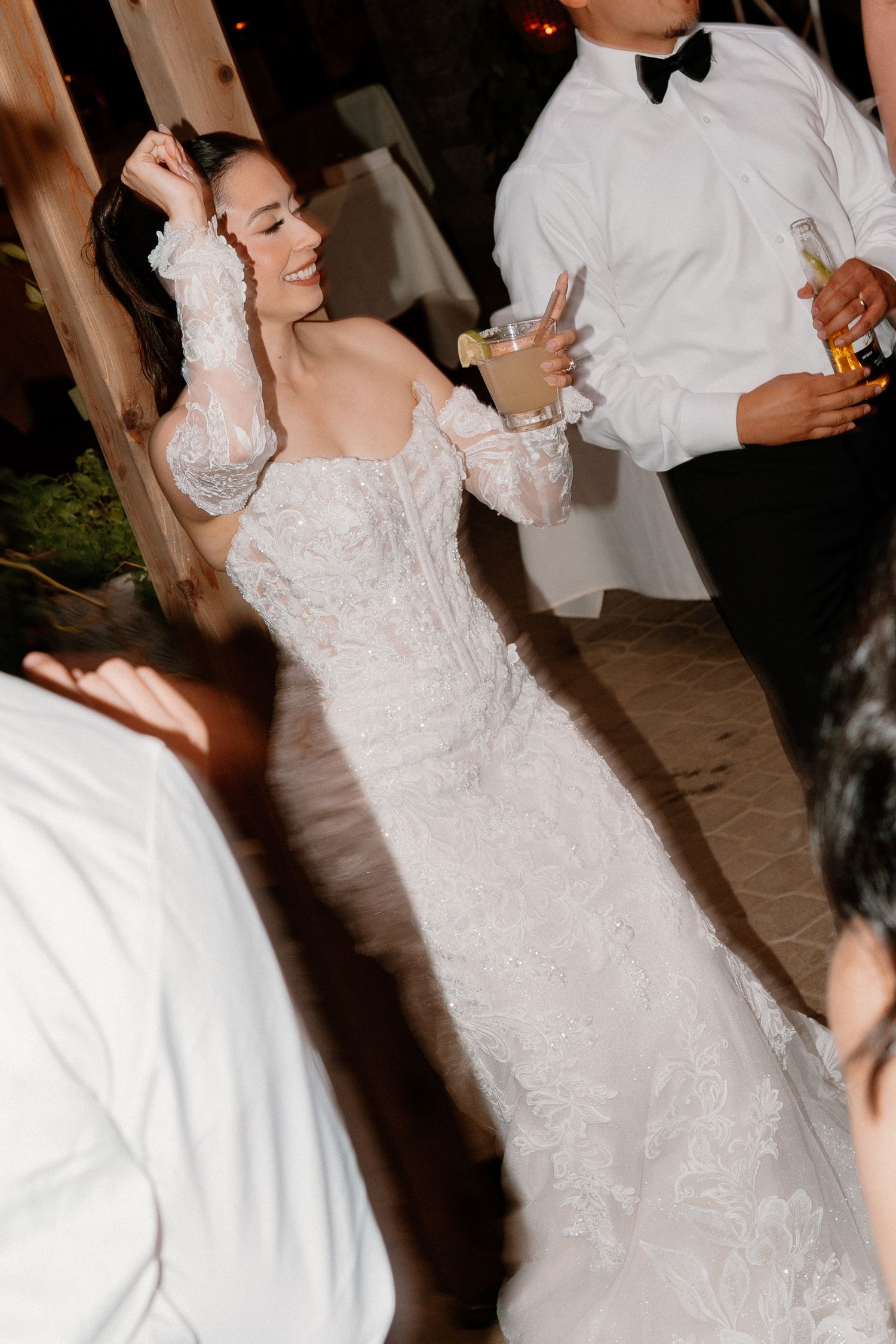 Radiant bride in off-shoulder lace gown dancing and holding a drink at her wedding reception.