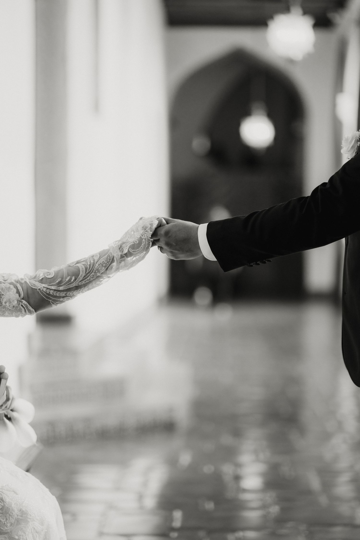 Black and white photo of a bride and groom holding hands through an arched hallway at their wedding venue.