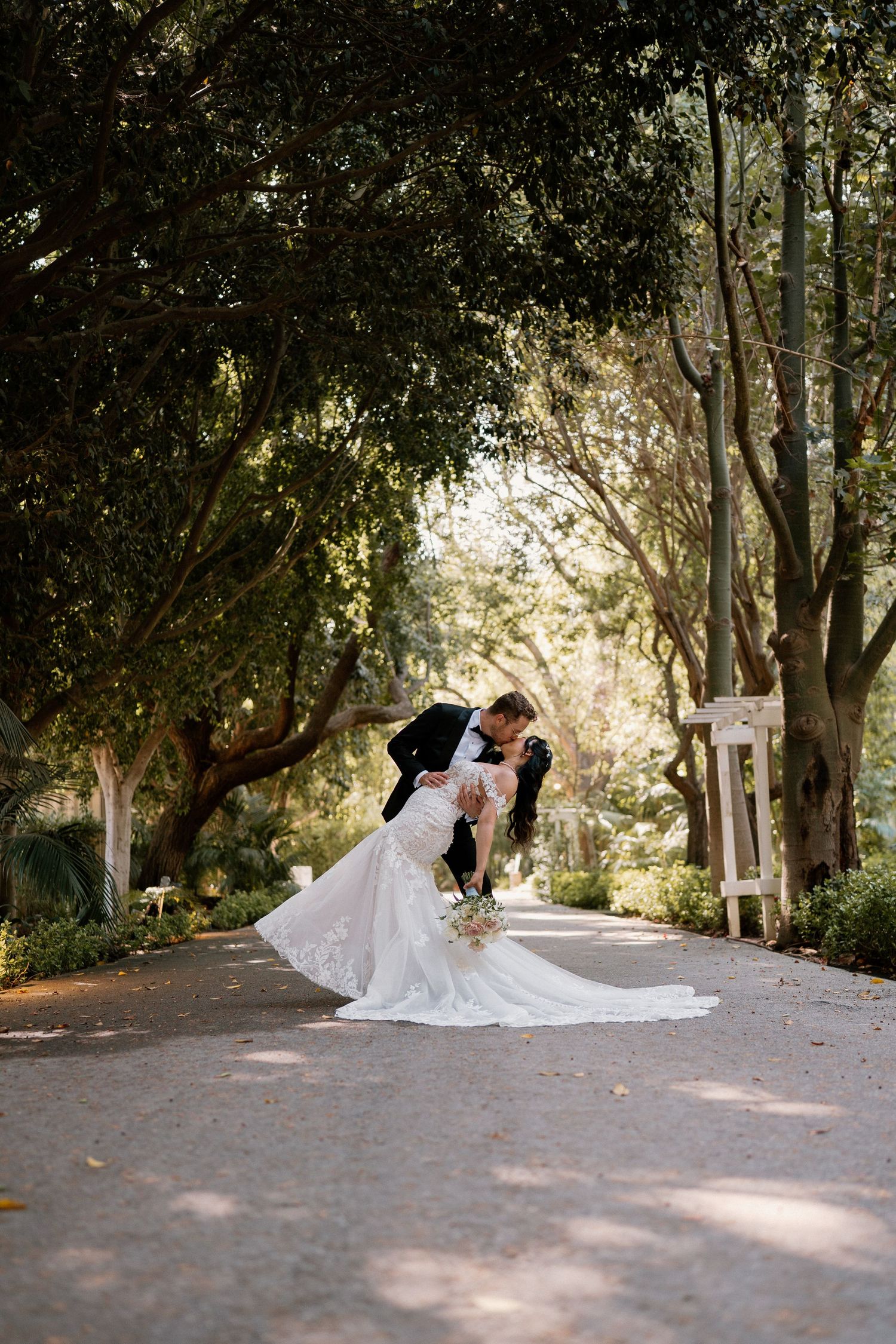 Groom dips bride for a romantic kiss on a tree-lined garden path in a stunning lace wedding gown.