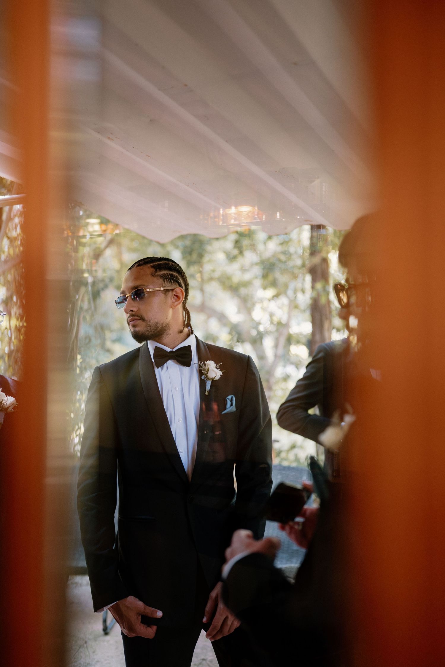Stylish groom in black tuxedo with bow tie and sunglasses waiting at wedding ceremony venue.