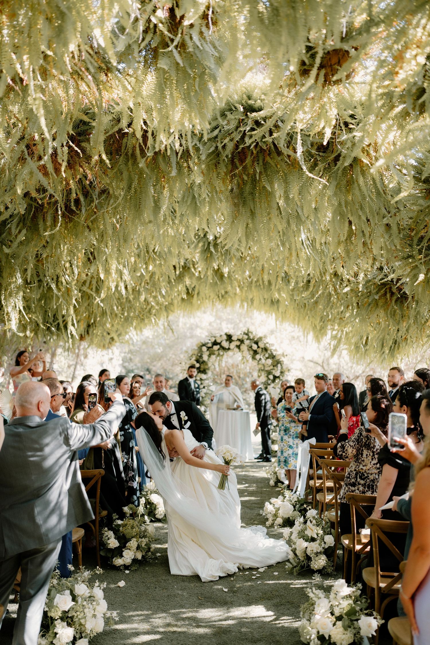 Wedding guests watch as a couple kisses at the altar under a stunning hanging greenery canopy.