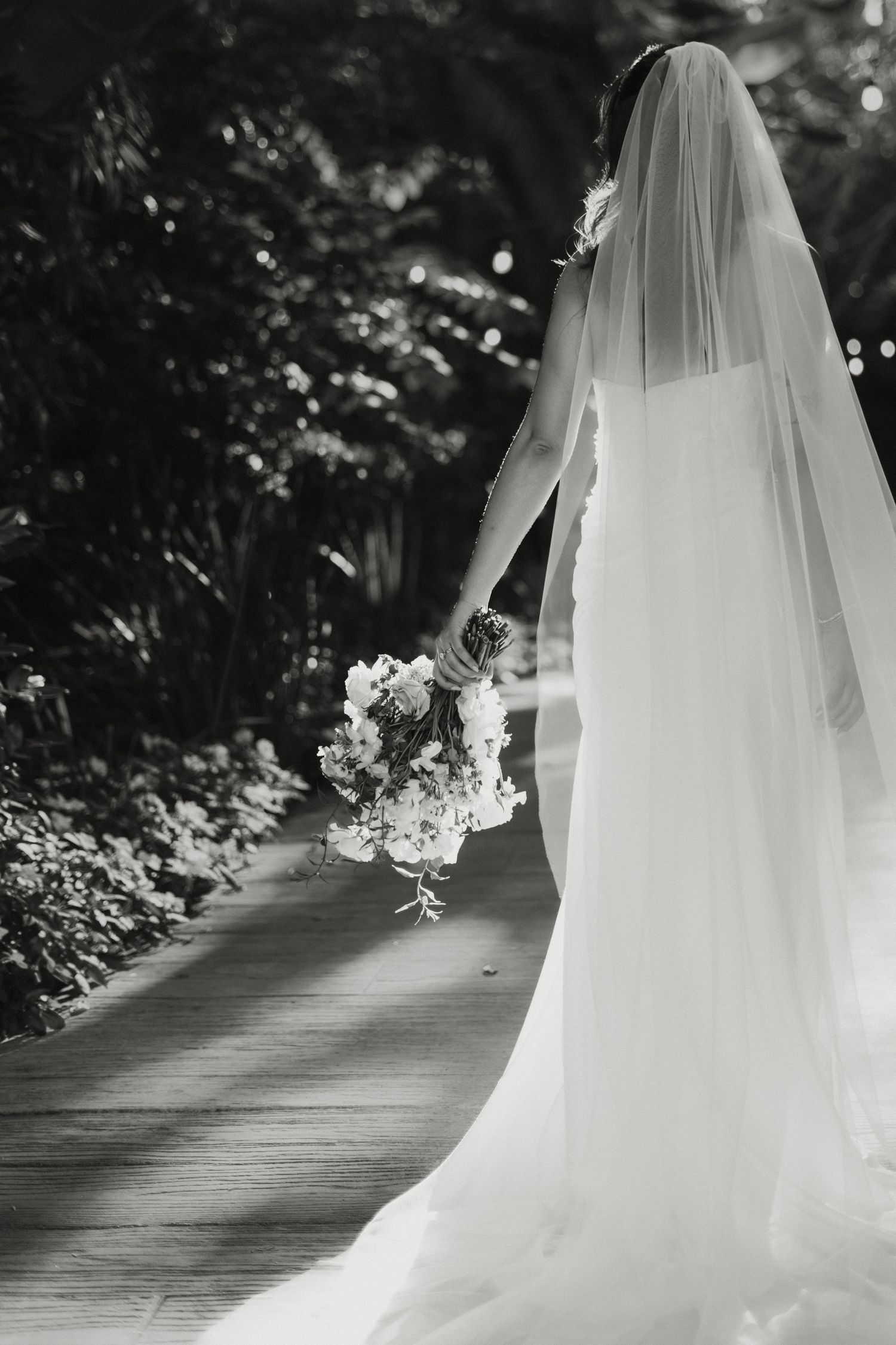 Black and white photo of bride walking garden path in flowing white gown and veil holding floral bouquet.