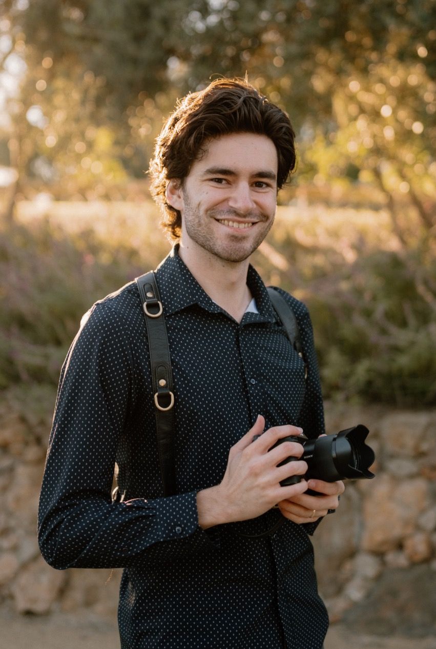 Smiling male wedding photographer holding camera with leather strap in golden hour sunlight outdoors.