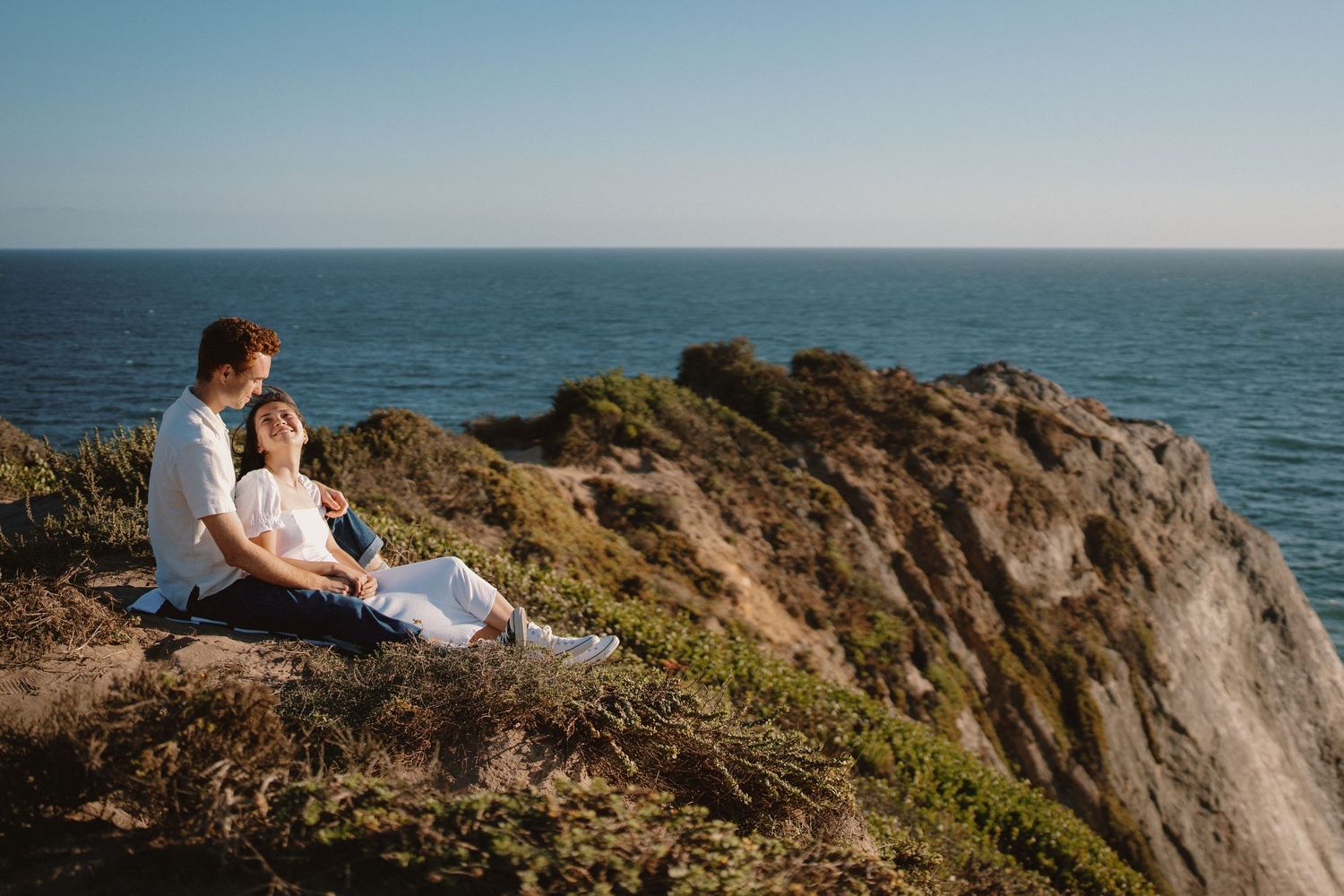 Romantic couple sitting on a coastal cliff overlooking the ocean on a sunny day.