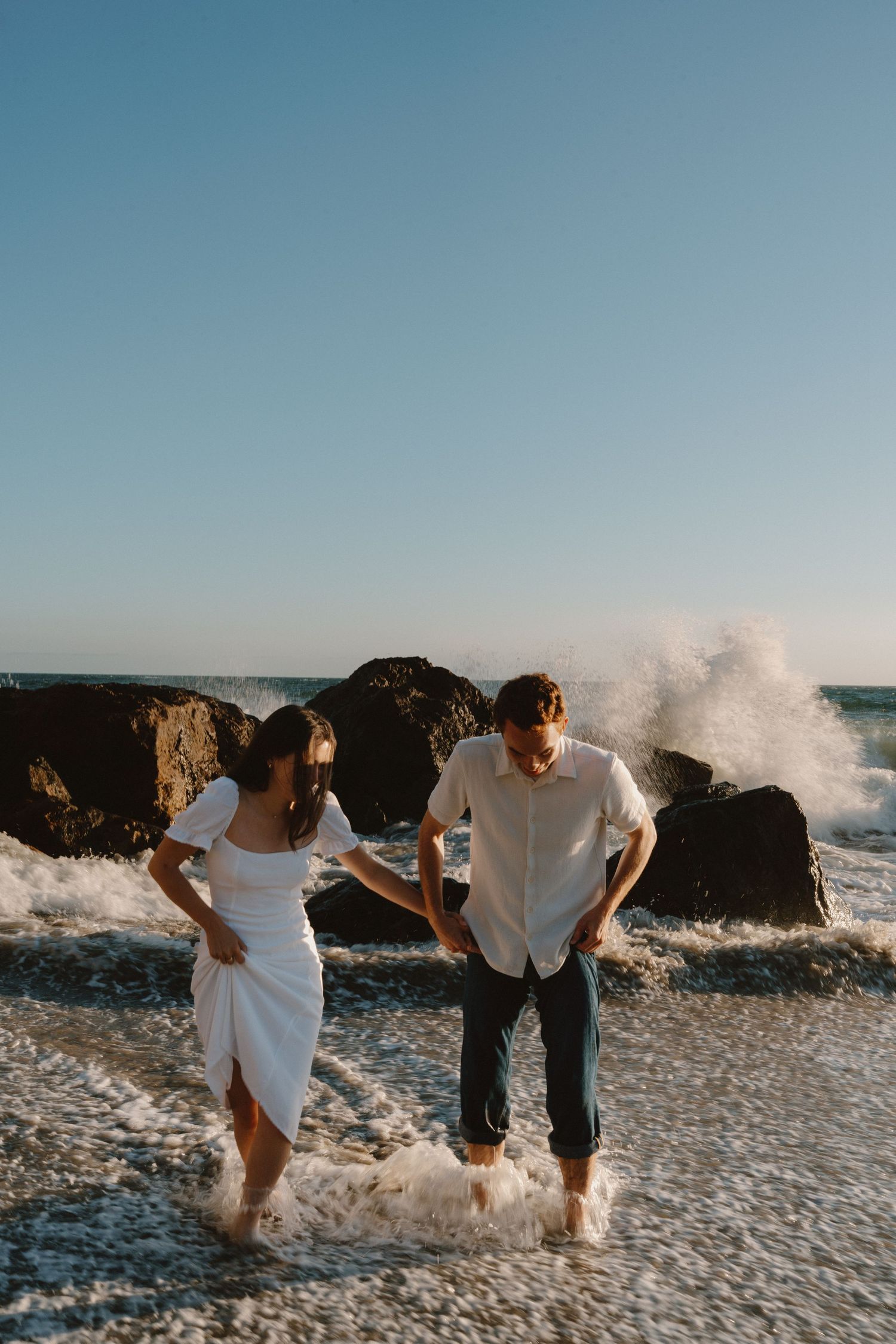 Couple laughing and holding hands while ocean waves crash around their feet on a rocky beach at golden hour.