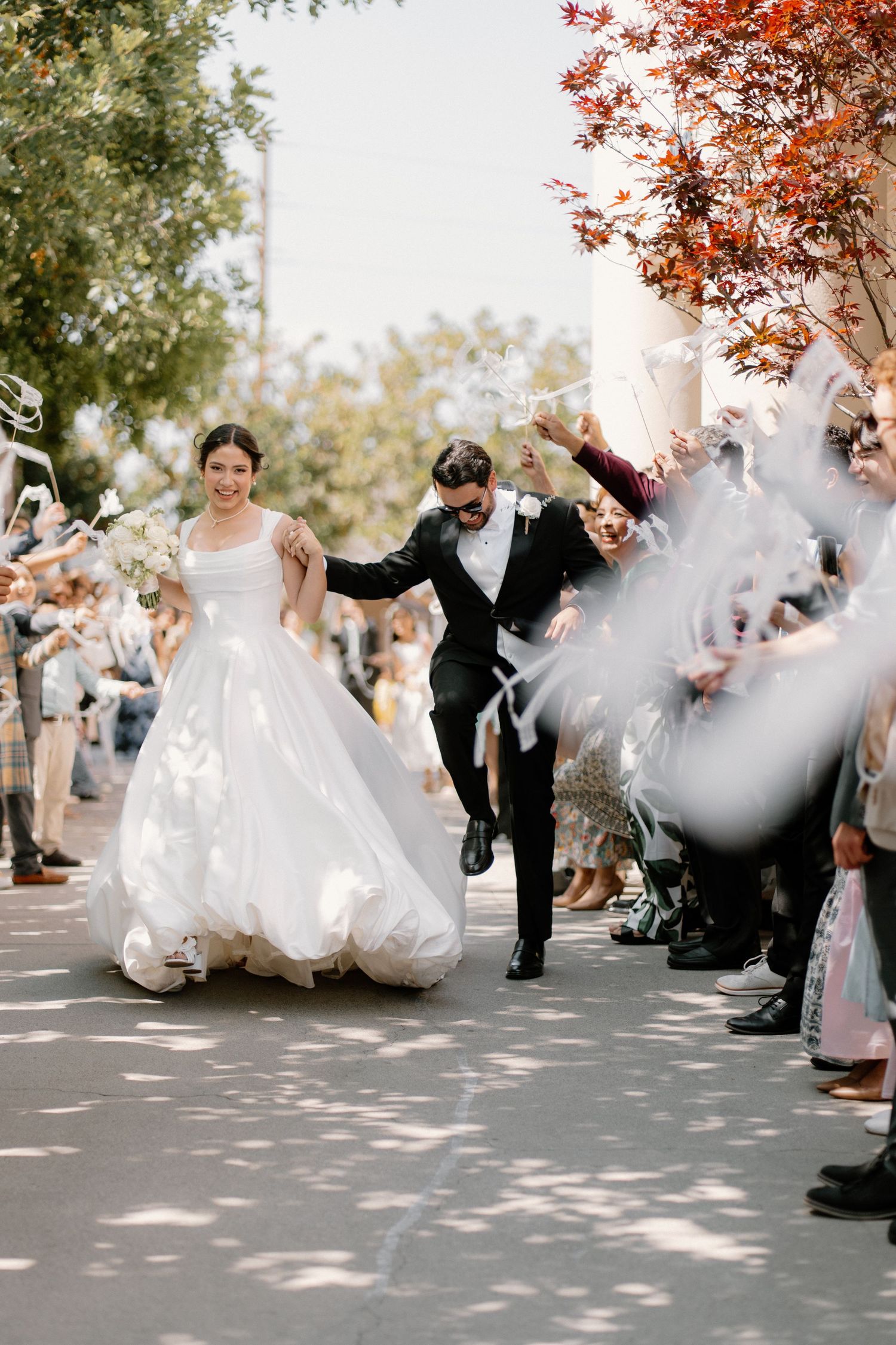Bride and groom run joyfully through ribbon wand send-off with cheering guests lining the sunny outdoor pathway.