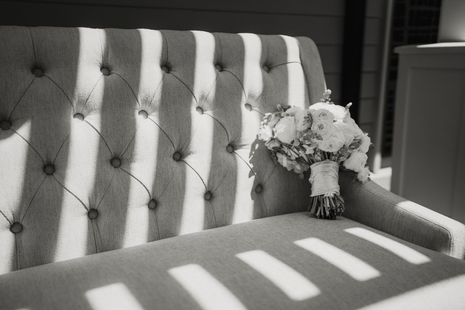 Black and white photo of a bridal bouquet resting on a tufted upholstered sofa with dramatic light and shadow.