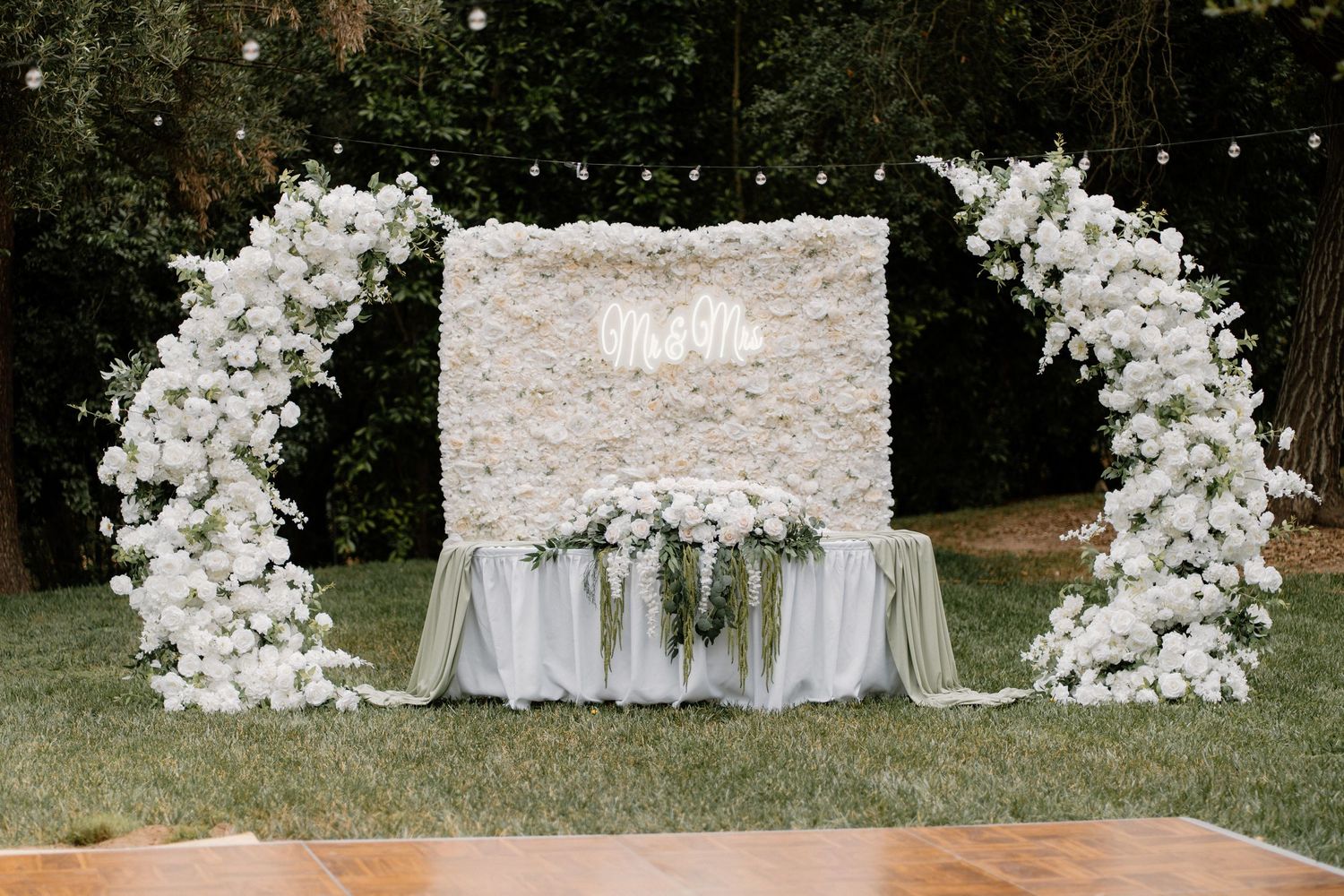 Elegant wedding backdrop with white flower wall, floral arches, and Mr. & Mrs. neon sign above a draped sweetheart table.