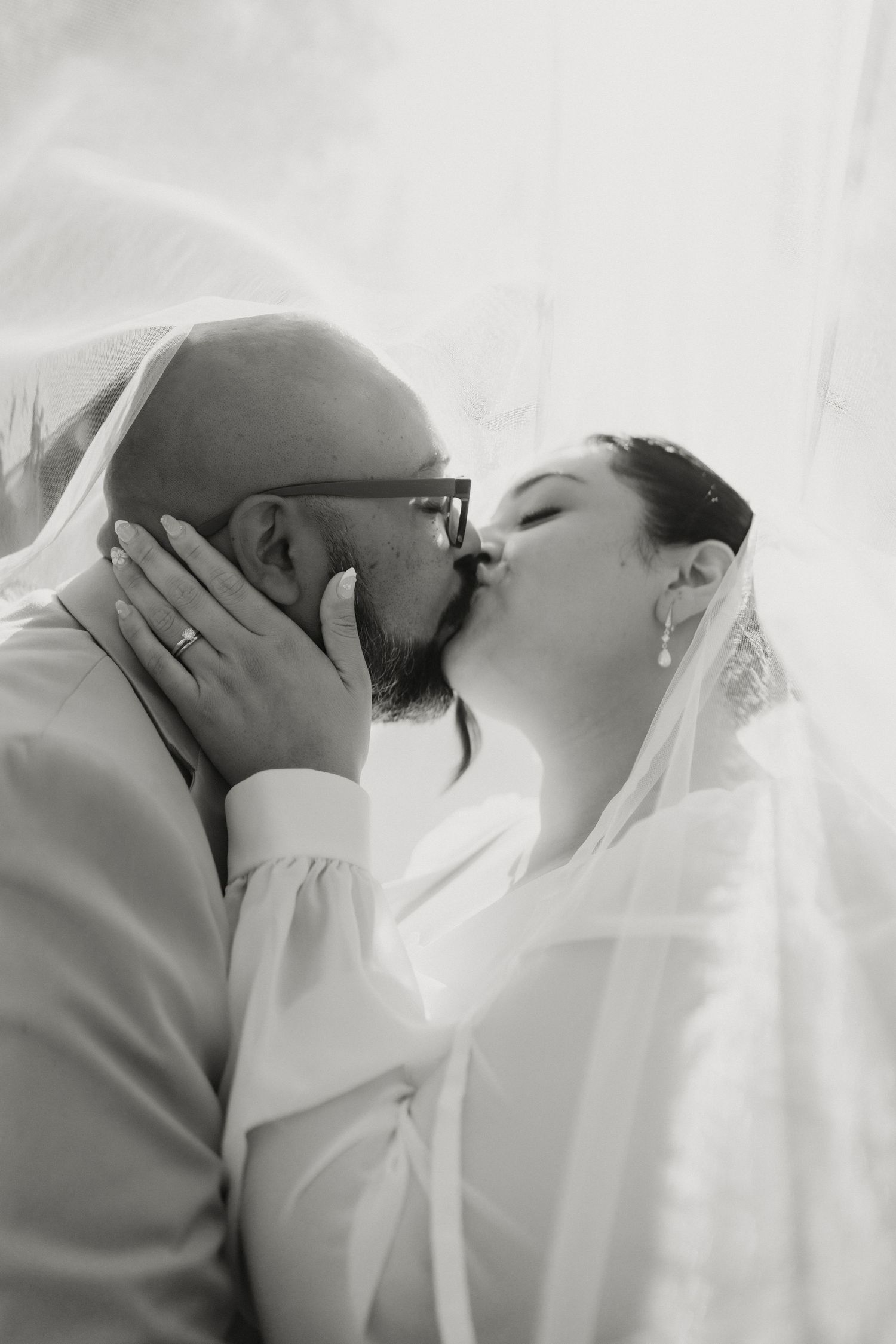 Black and white romantic photo of bride and groom kissing under flowing wedding veil.