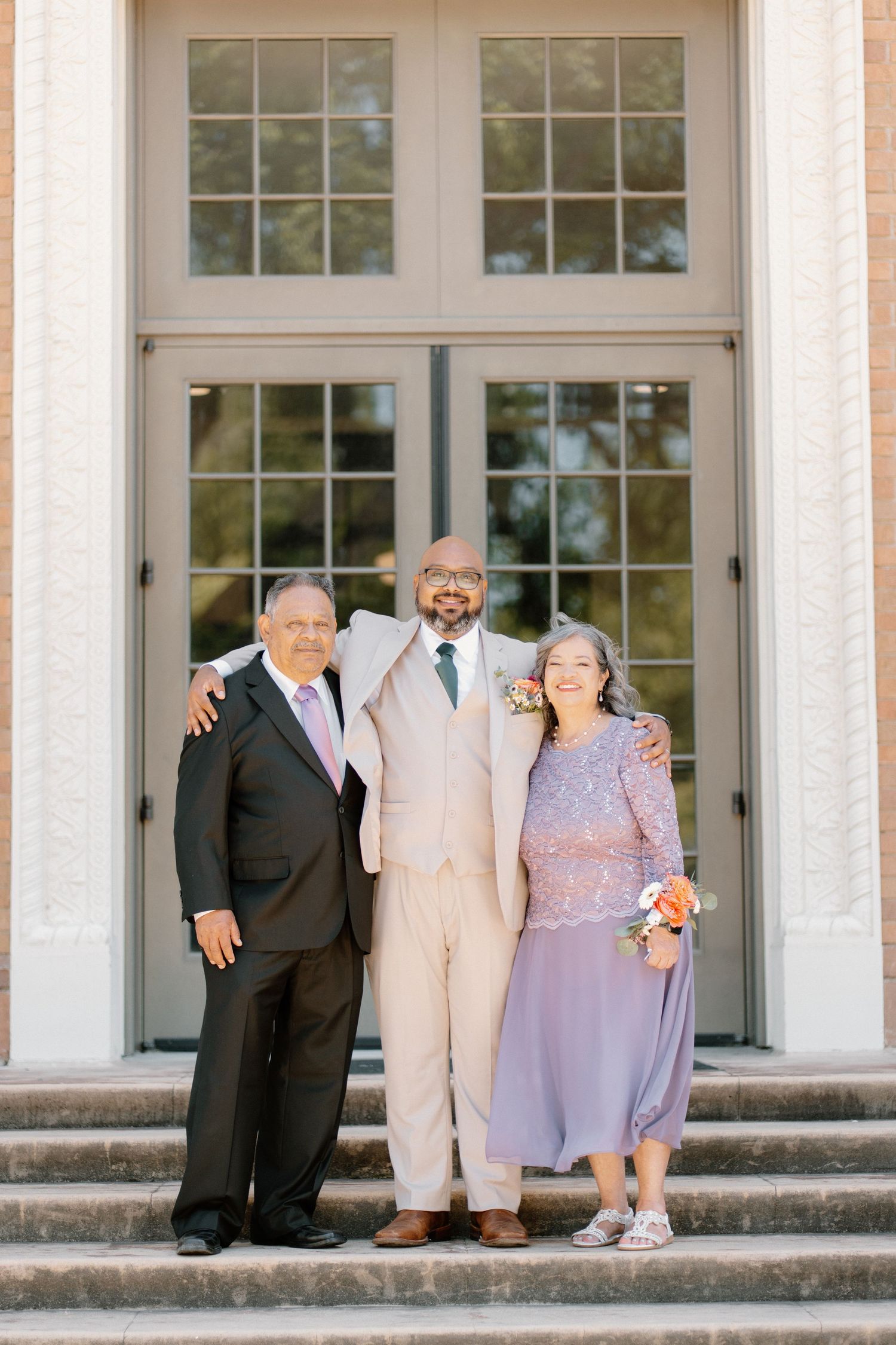 Groom in tan suit poses with parents on church steps at wedding ceremony.
