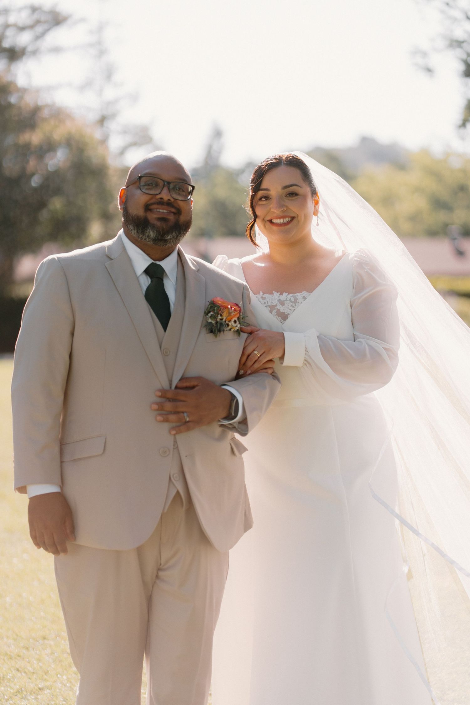 Bride and groom smiling together outdoors, bride in white gown with veil, groom in tan suit.
