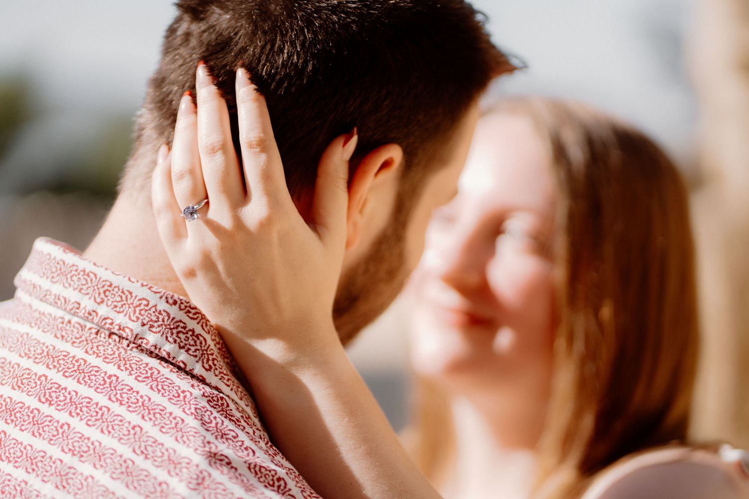 Couple sharing a romantic moment, woman smiling with engagement ring visible on hand resting on mans shoulder.