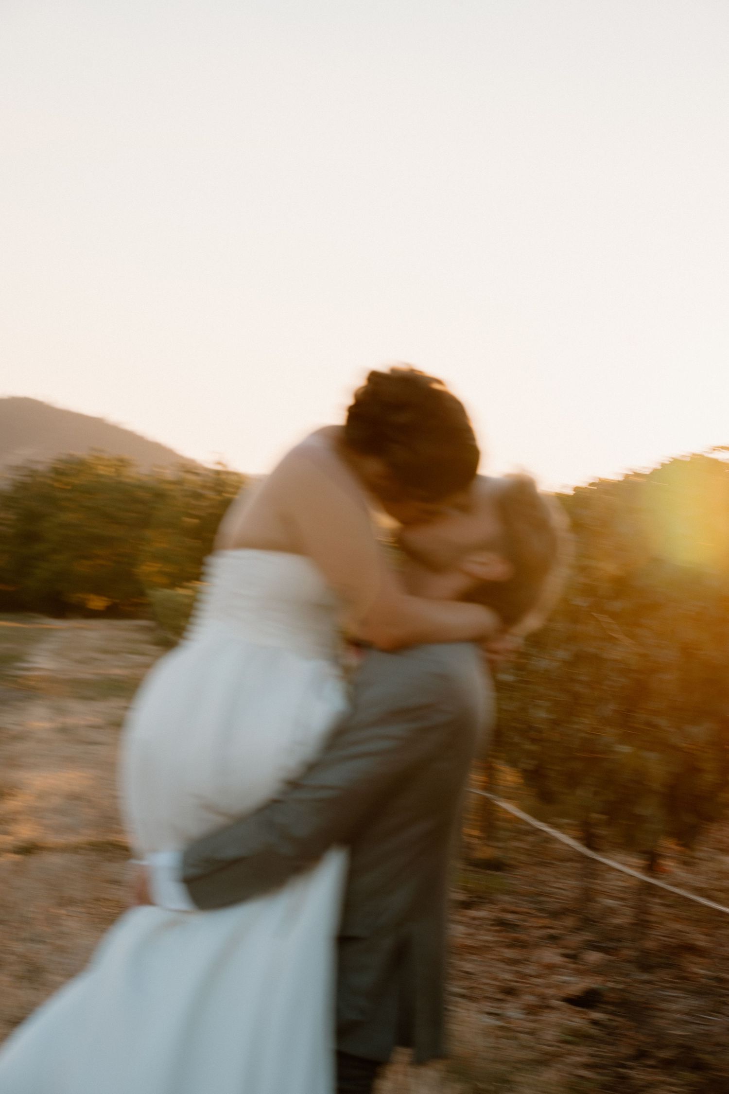 Blurry romantic wedding couple kissing at golden sunset in vineyard, bride in white gown embraced by groom.