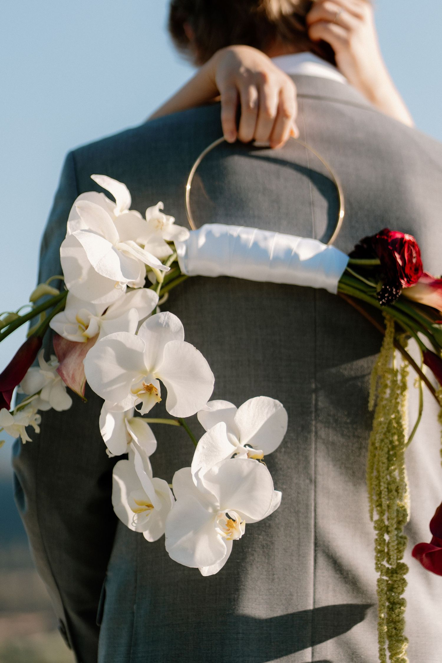 Bride holding floral hoop with white orchids and red flowers while embracing groom in gray suit.