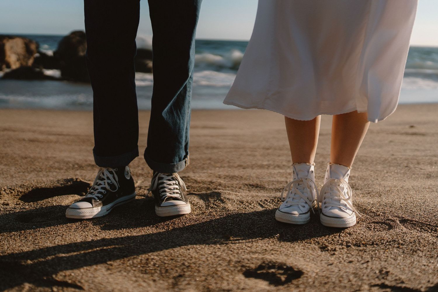 Close-up of couple's feet wearing matching Converse sneakers standing on sandy beach near ocean waves.