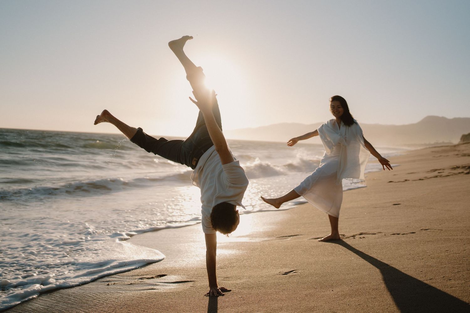 Man doing cartwheel on beach at sunset while woman watches nearby.