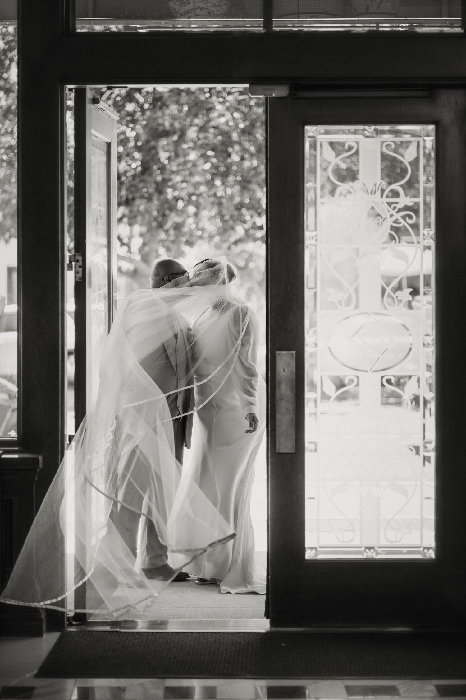 Black and white photo of bride and groom kissing in doorway, wedding veil flowing gracefully around them.