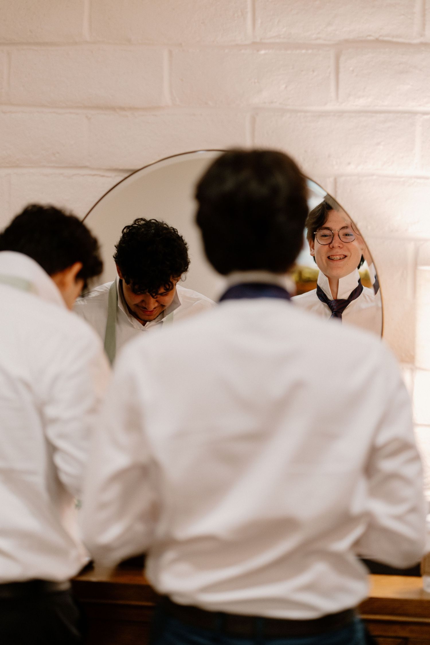 Groomsmen in white dress shirts help each other get ready while smiling in a round mirror.