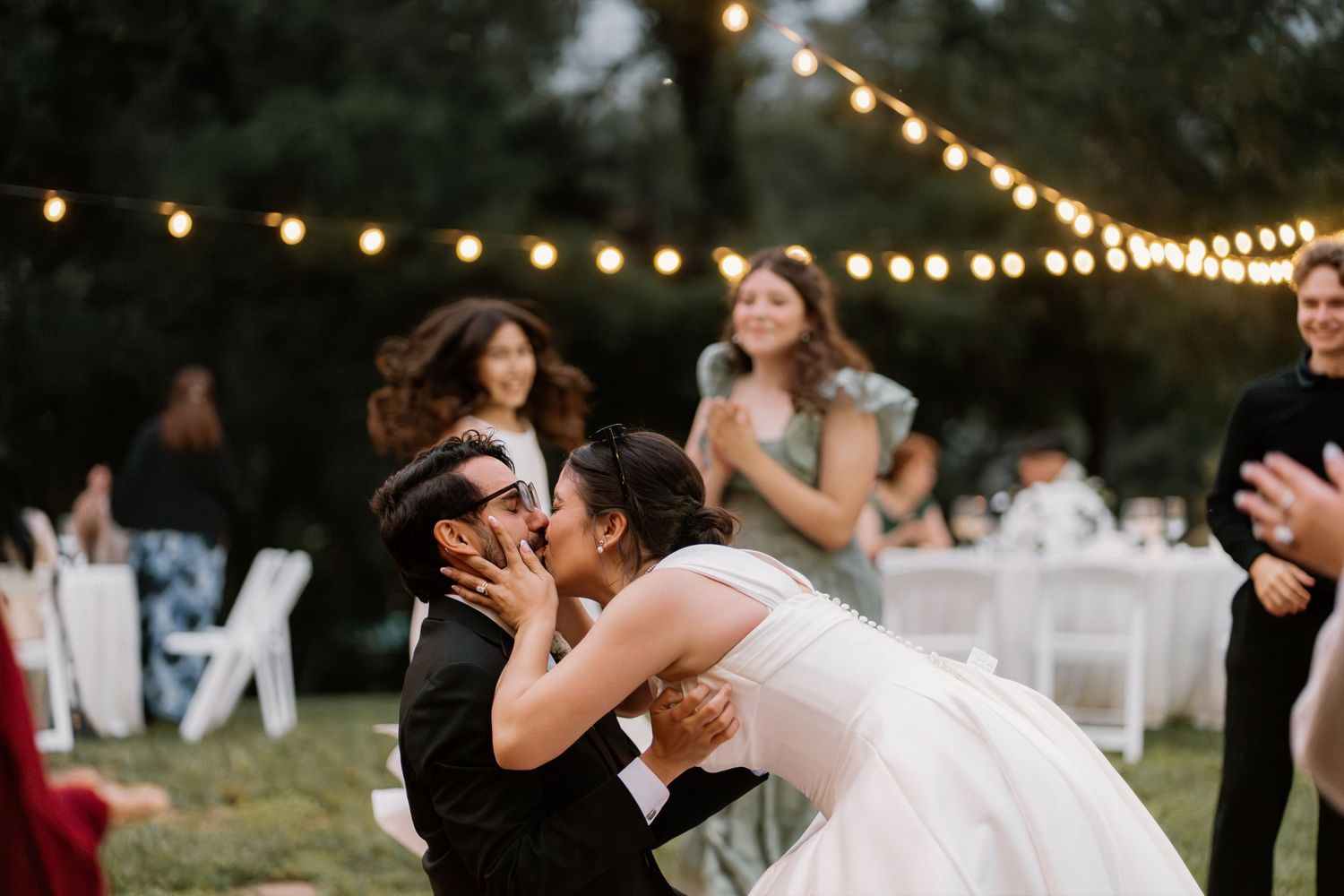 Bride and groom share a romantic kiss during outdoor wedding reception with string lights and guests in background.