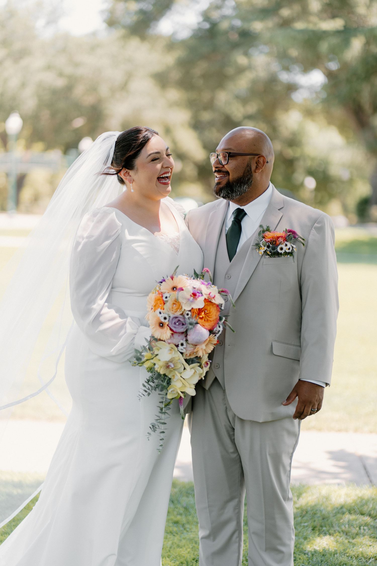 Smiling bride in white gown holding colorful bouquet stands with groom in gray suit outdoors on wedding day.