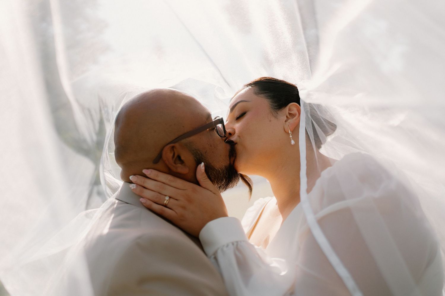 Romantic newlyweds share a tender kiss beneath a flowing white bridal veil in soft light.