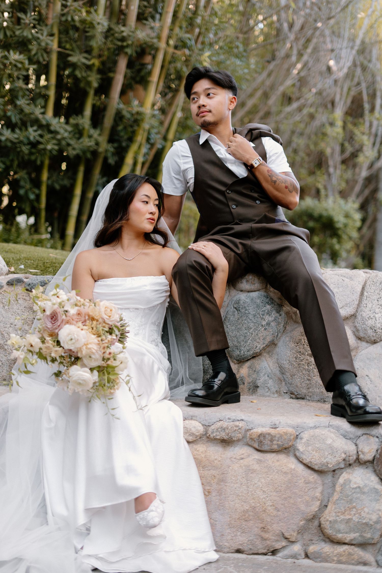 Bride and groom pose on stone steps in garden setting, bride holding soft pink and white floral bouquet.