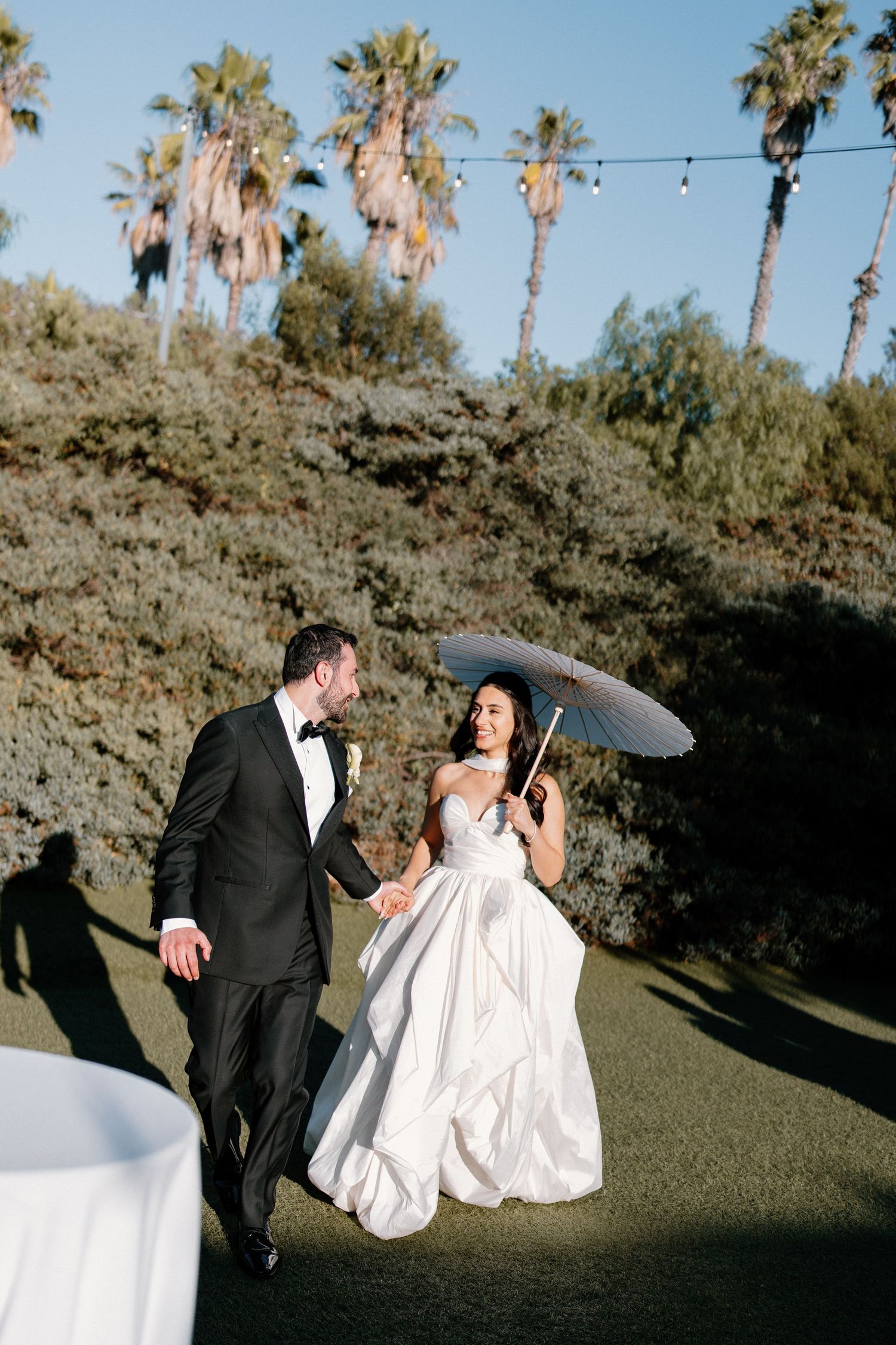 A smiling bride holding a parasol walks hand-in-hand with her groom among tall palm trees.