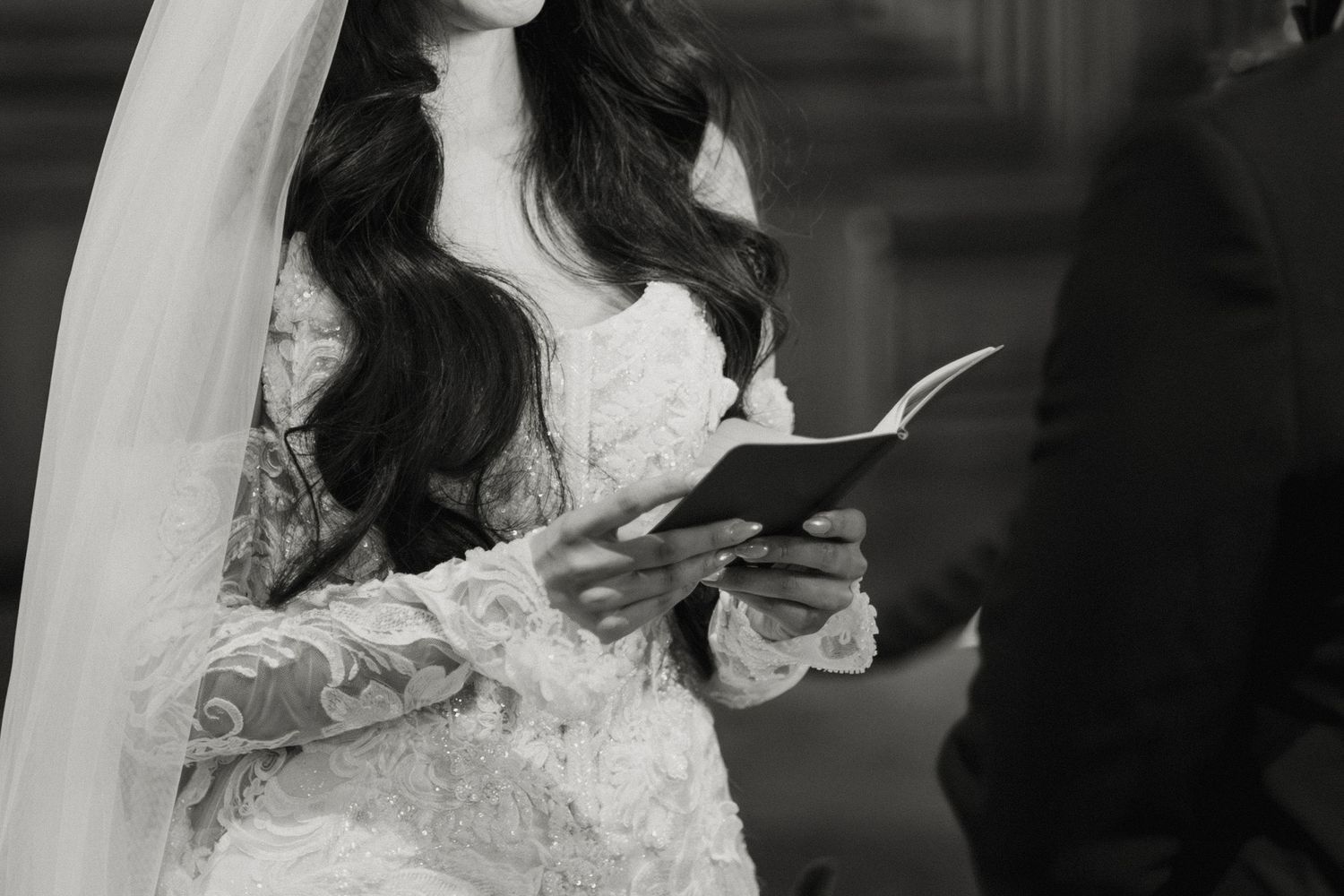 Bride in lace wedding gown and veil reading vows from a book during wedding ceremony in black and white.